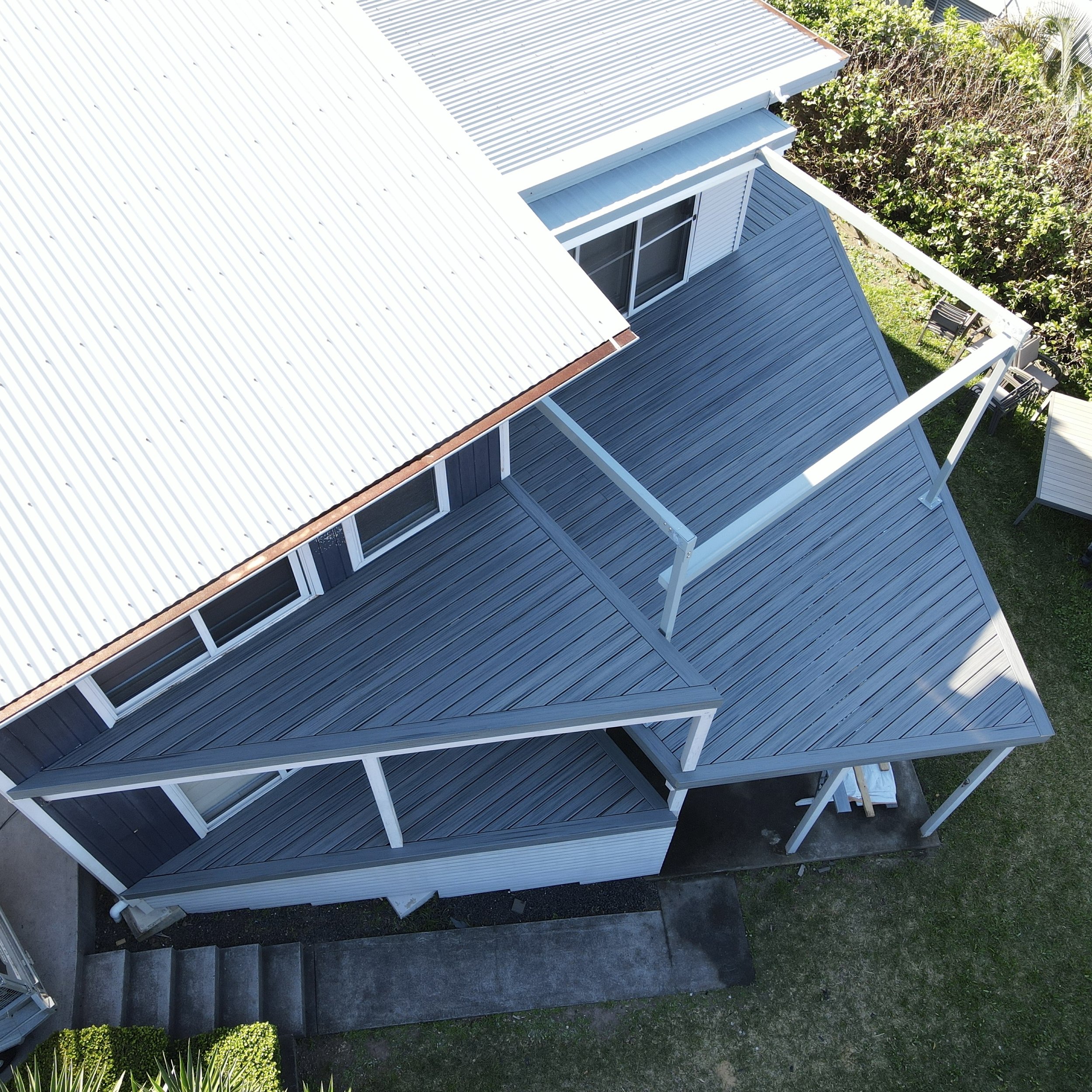 A house with a white corrugated metal roof, blue siding, and large sliding glass doors, surrounded by a green garden with trees and bushes.