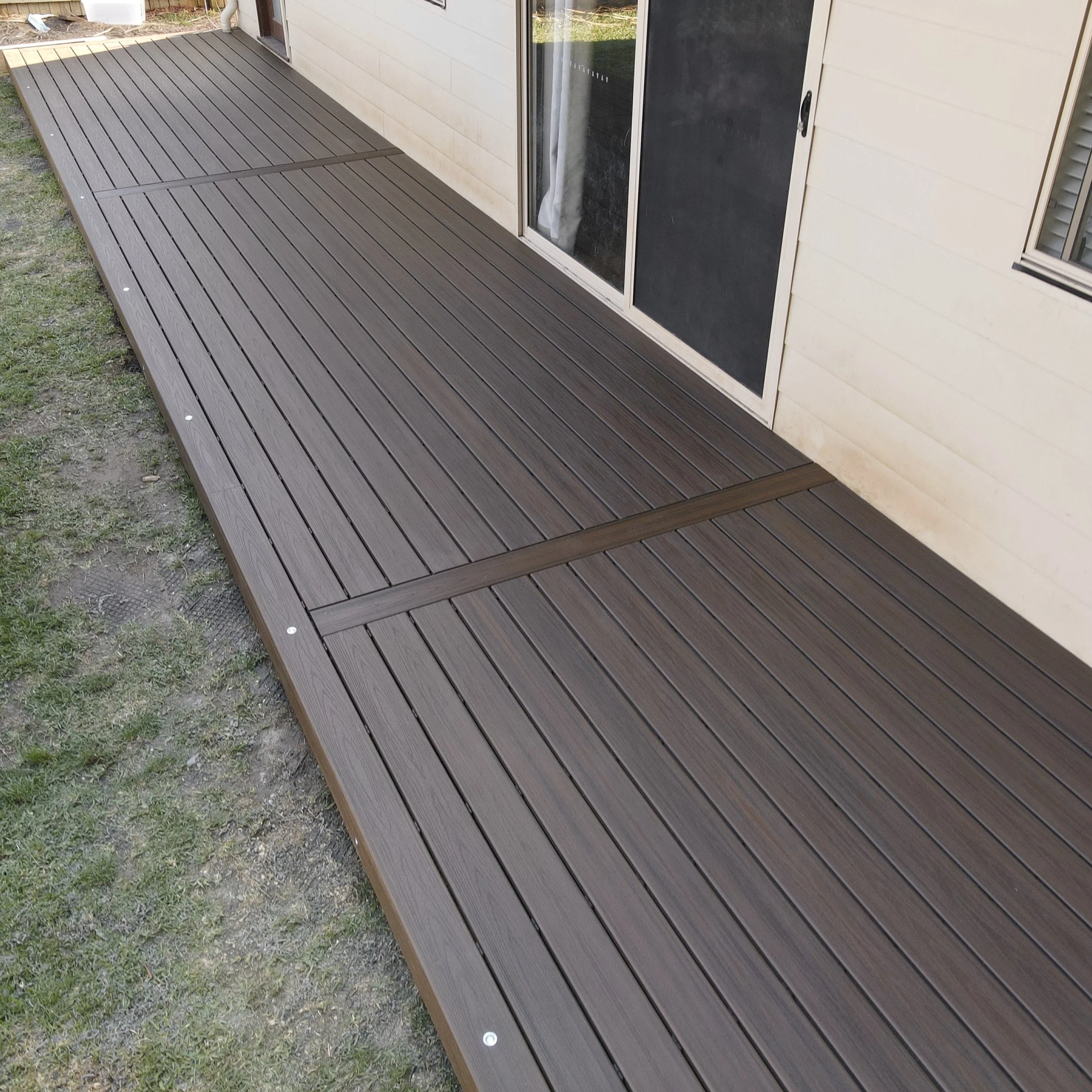 A brown composite deck running the length of the house with a sliding glass door and a screen door, surrounded by grass and dirt. Built and installed on the Central Coast by Platinum Decks.