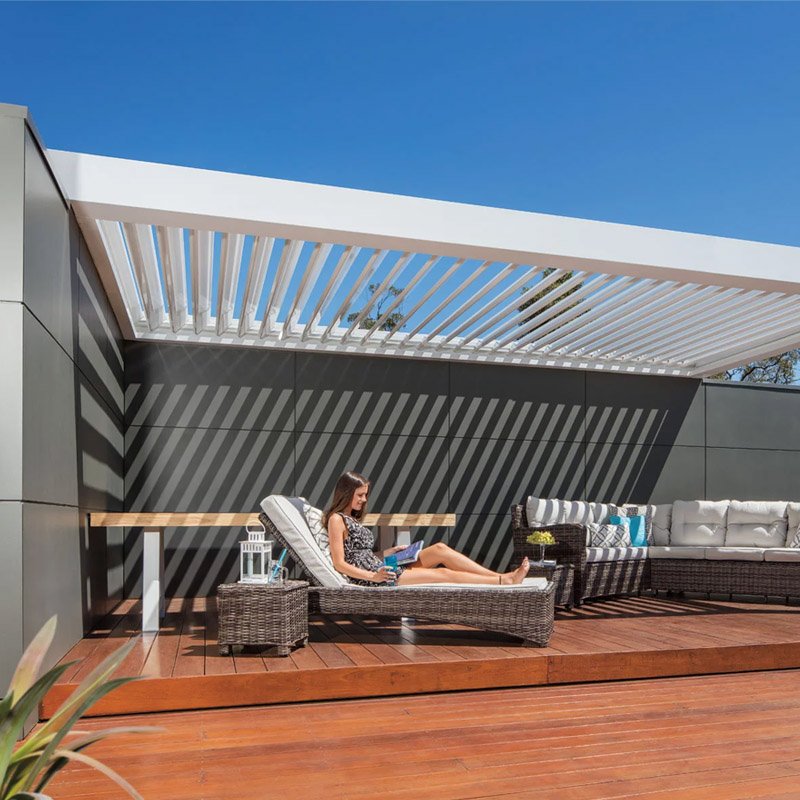 Woman relaxing on a lounge chair on a rooftop patio, called a deck, with modern furniture, under a white pergola casting shadows on the wall, against a clear blue sky.