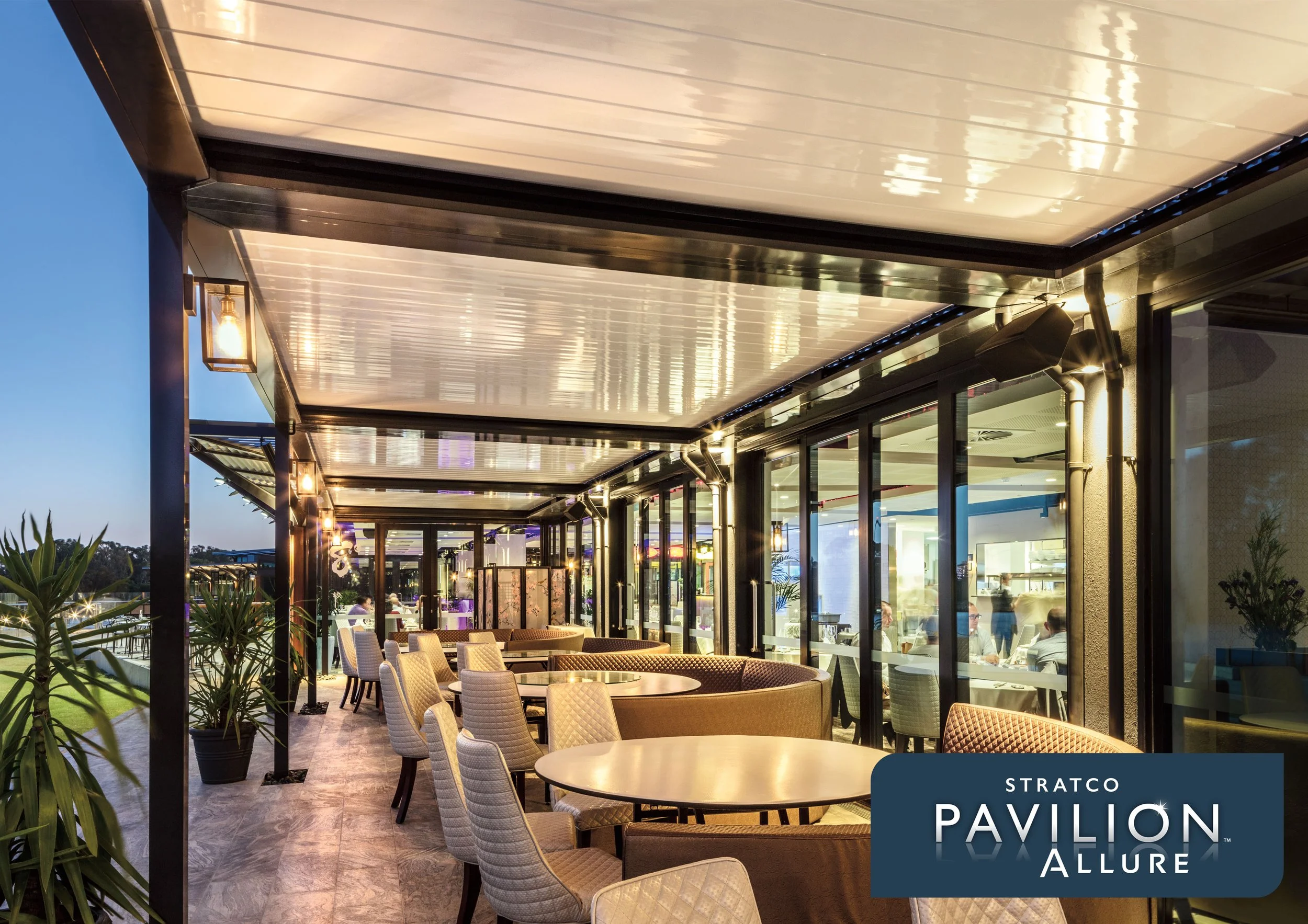 The image shows a modern outdoor dining area at dusk with round tables and beige chairs, covered by a metallic patio with built-in lighting, adjacent to a building with large glass windows and potted plants. Platinum Decks authorised installer.
