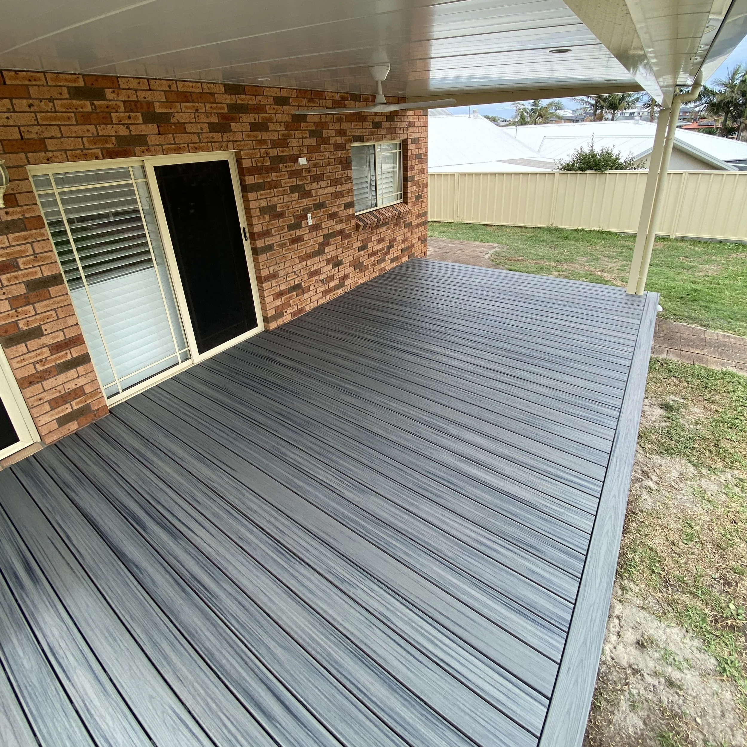 Backyard patio with wooden deck, brick house wall, sliding glass door, small window, ceiling fan, and a white fence. In the background, there are palm trees and neighboring houses with roofs.