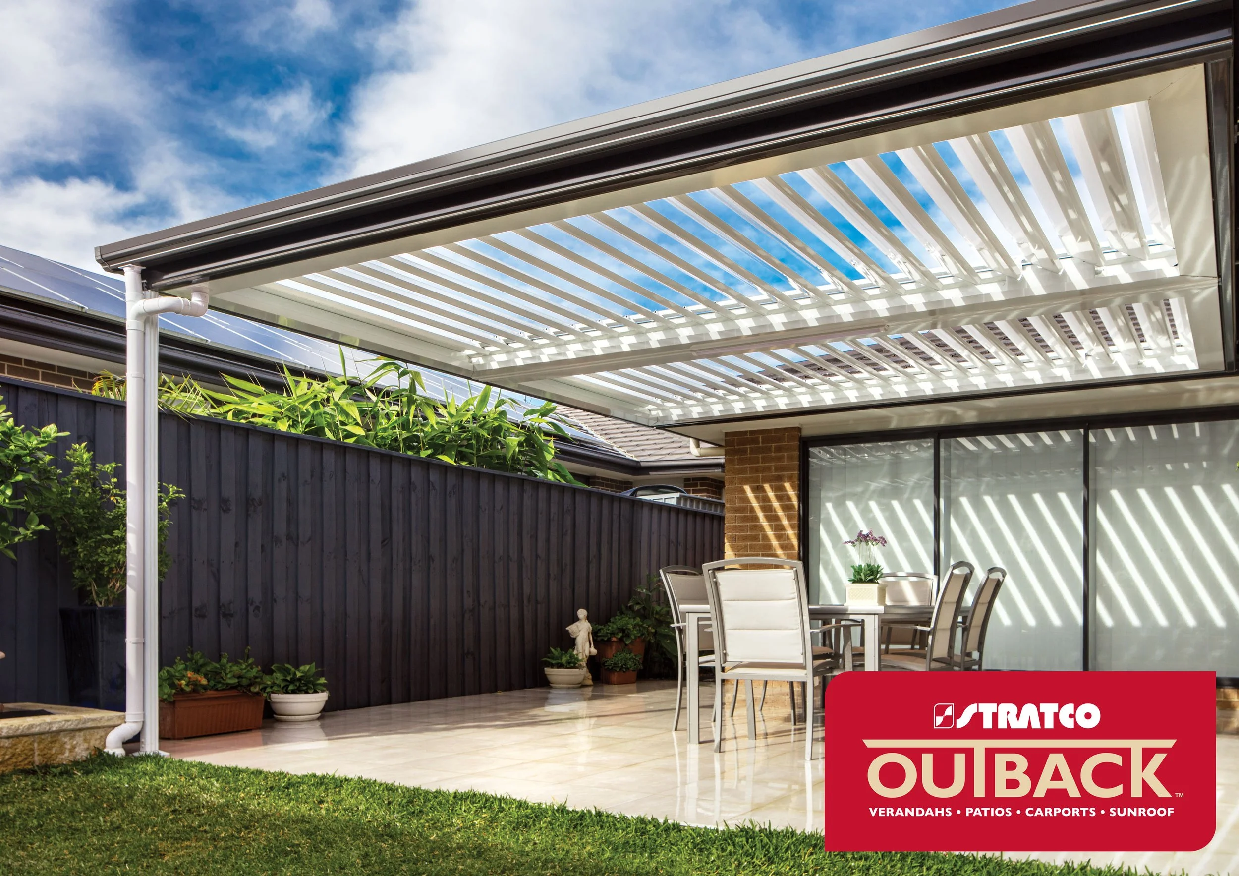 A backyard patio with a clear roof structure, outdoor dining table with chairs, potted plants, a fenced yard, and a house with visible solar panels on the roof. Platinum Decks authorised installer.