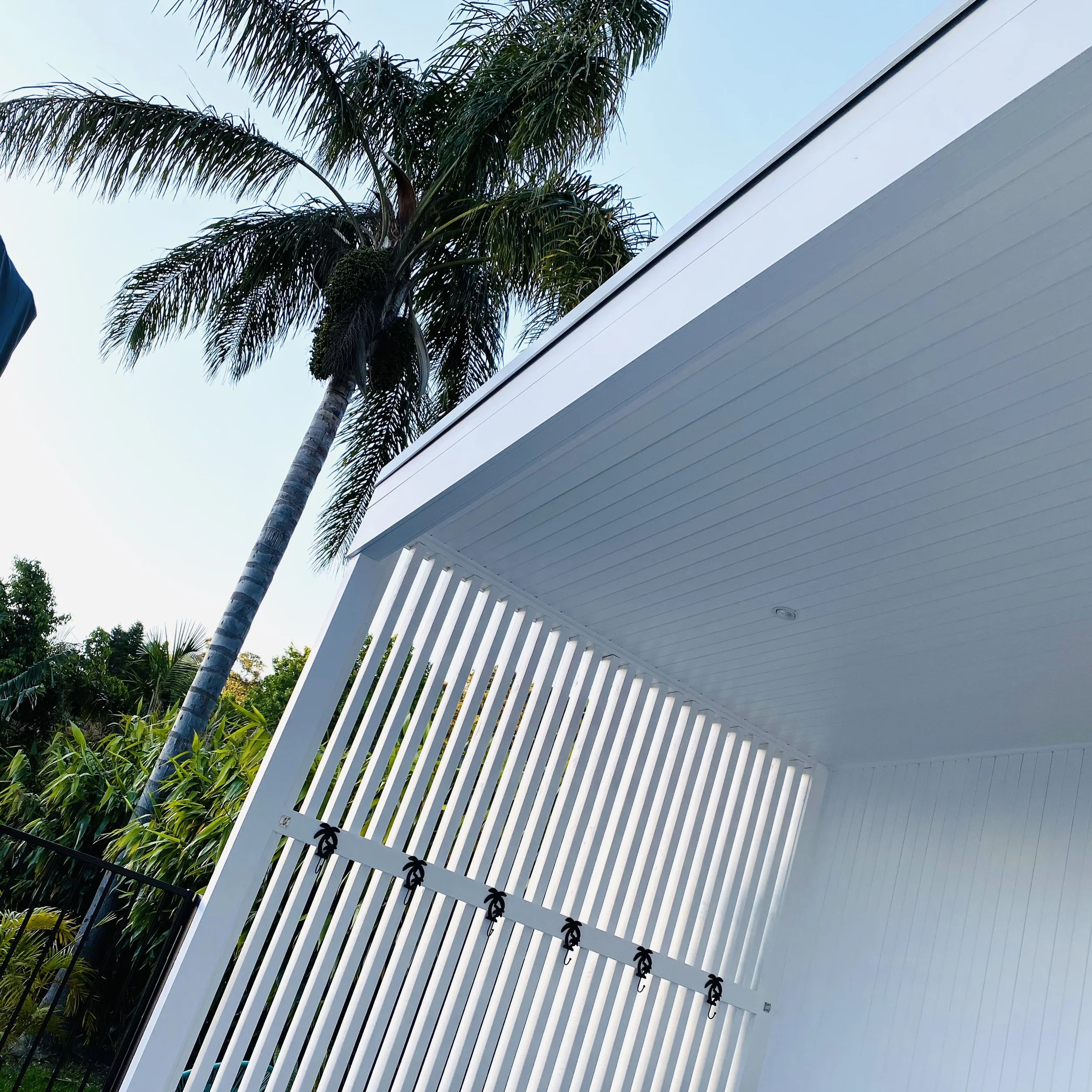 A white pool house with vertical privacy screen with a palm tree next to the buildingg and a clear blue sky in the background. Built, designed and installed by Platinum Decks