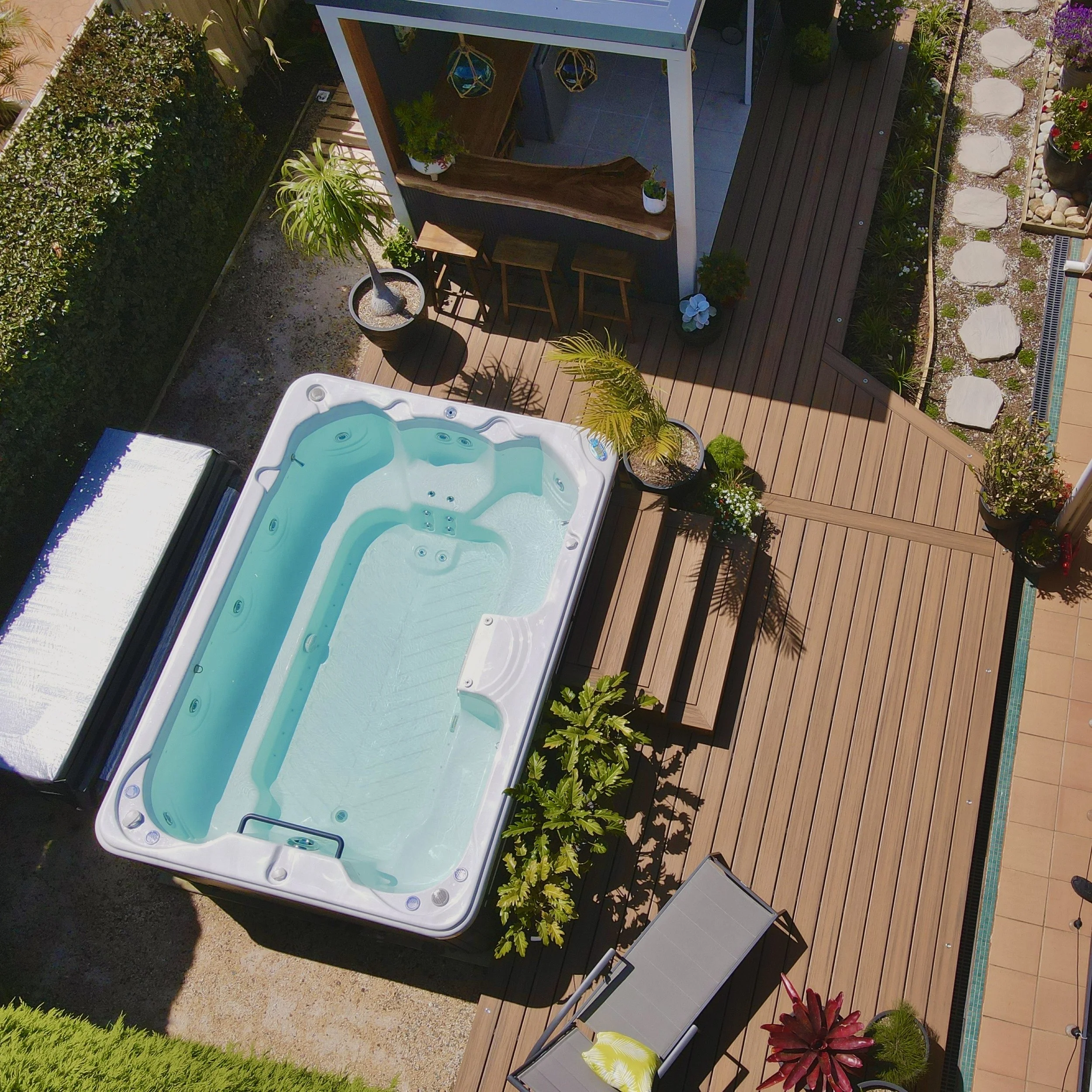 Aerial view of a backyard patio built by Platinum Decks with a brown ncomposite deck,  spa, green potted plants and garden beds filled with stones and colourful flowers.