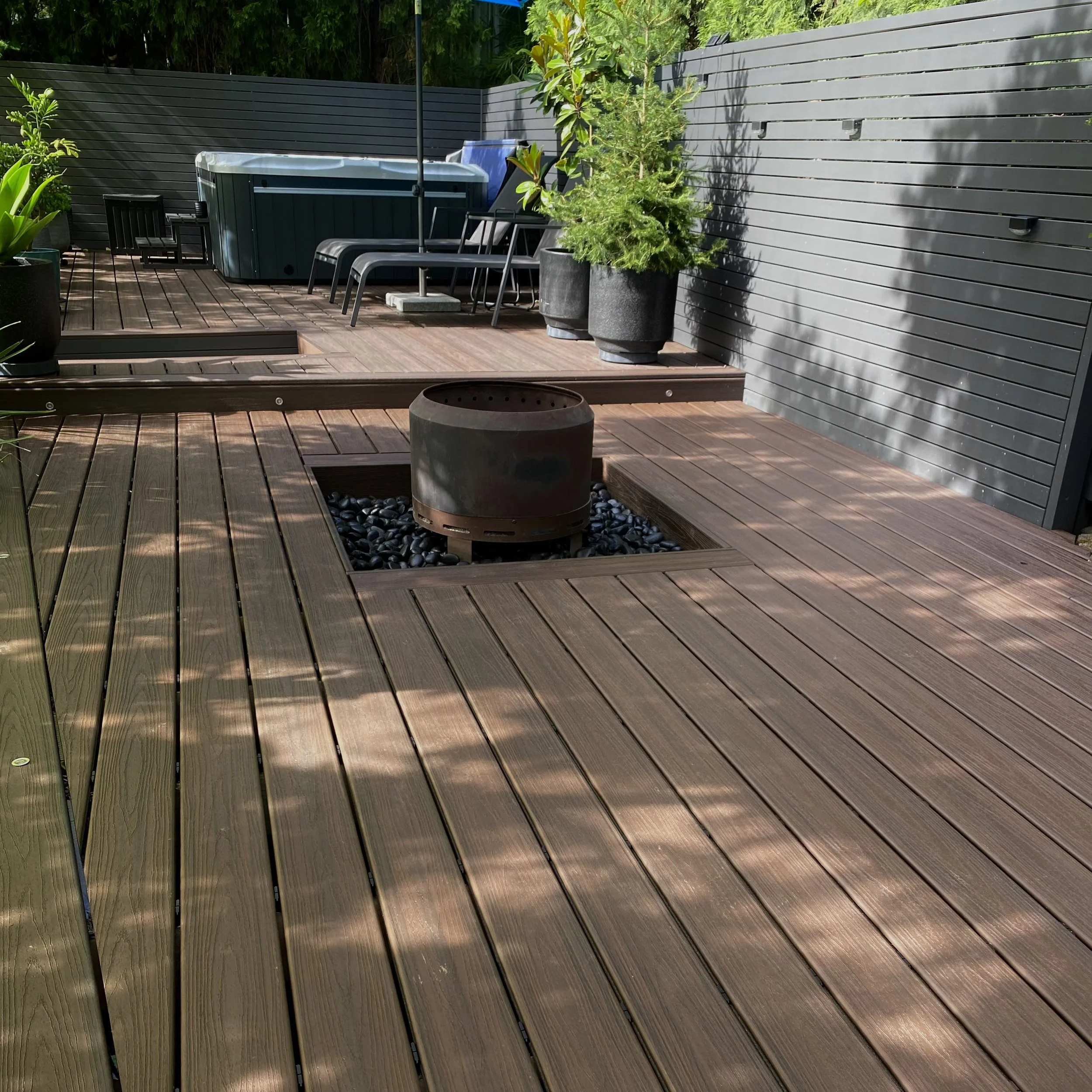 A backyard modern composite low maintenance deck area with potted plants, black chairs, a spa, and tall grey fencing surrounded by greenery.