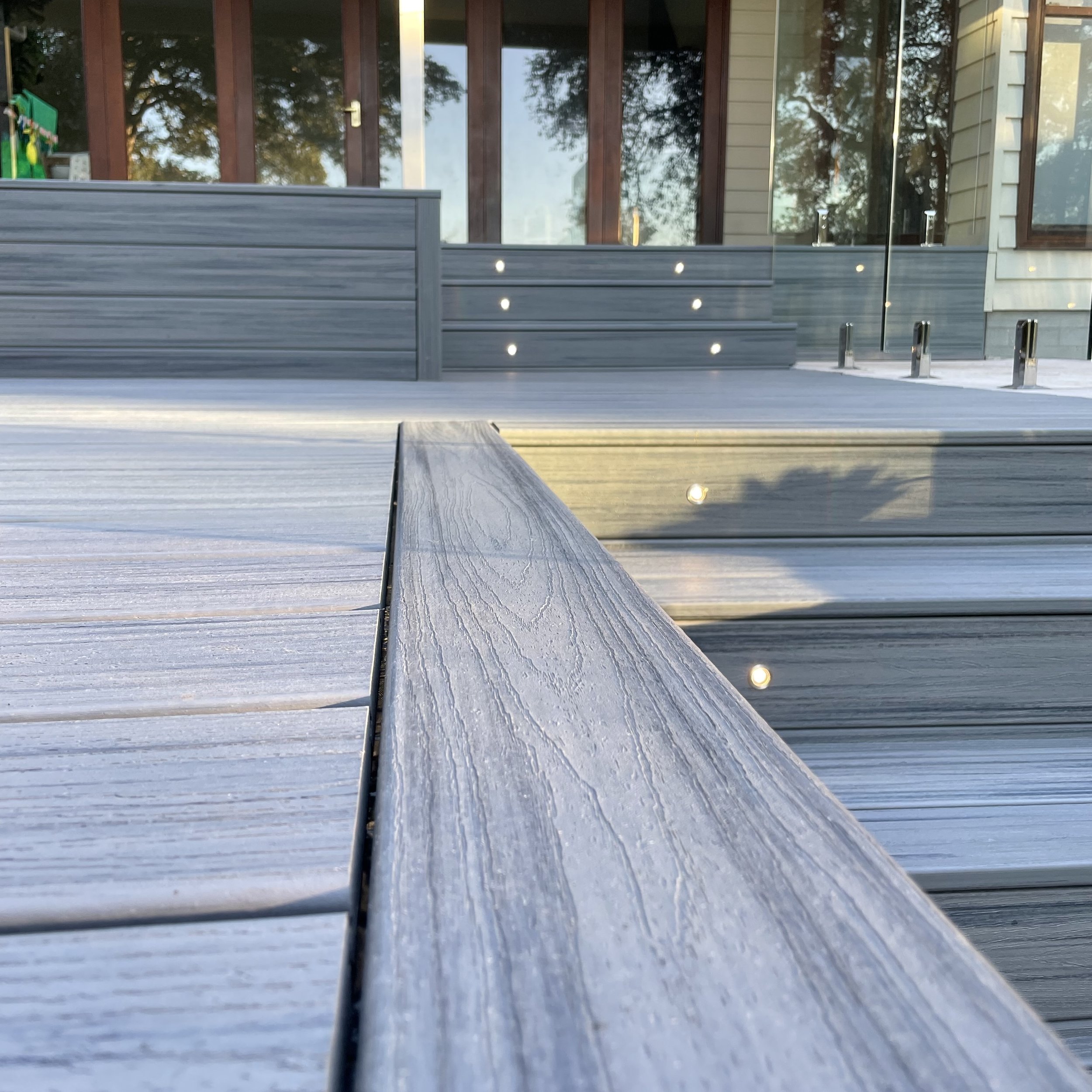 Close-up of composite wooden deck flooring with horizontal planks and small circular embedded lights, with a glass balstrade in the background.