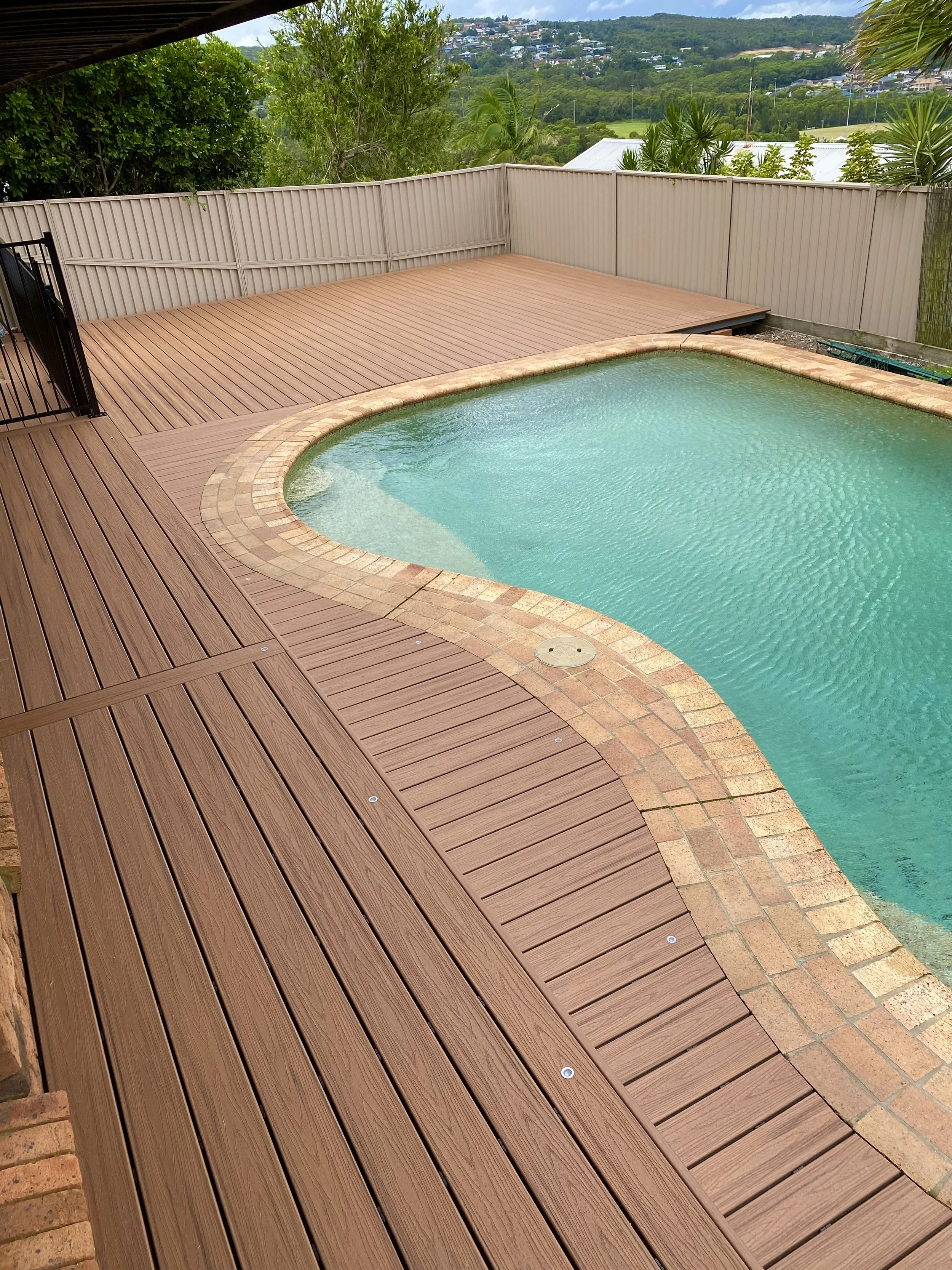 A reddish brown composite deck curving neatly around a pool with tropical plants and a flowing layout, installed by Platinum Decks for a relaxed resort‑style feel