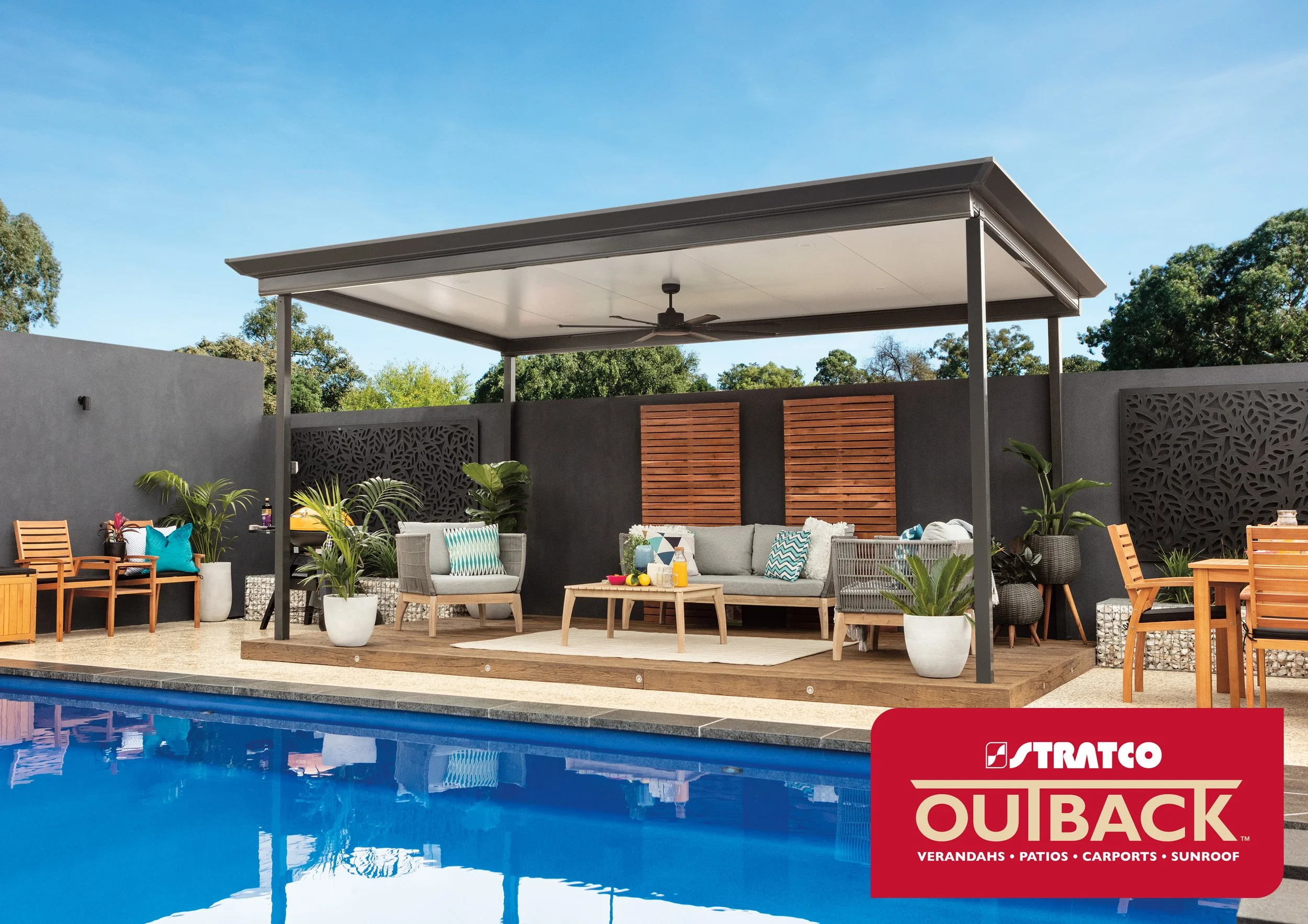 A modern outdoor living area with a pool Stratco roof,  sitting and dining furniture, a shade structure with ceiling fans, potted plants, and decorative wooden panels on a dark wall, under a clear blue sky. Platinum Decks authorised installer.