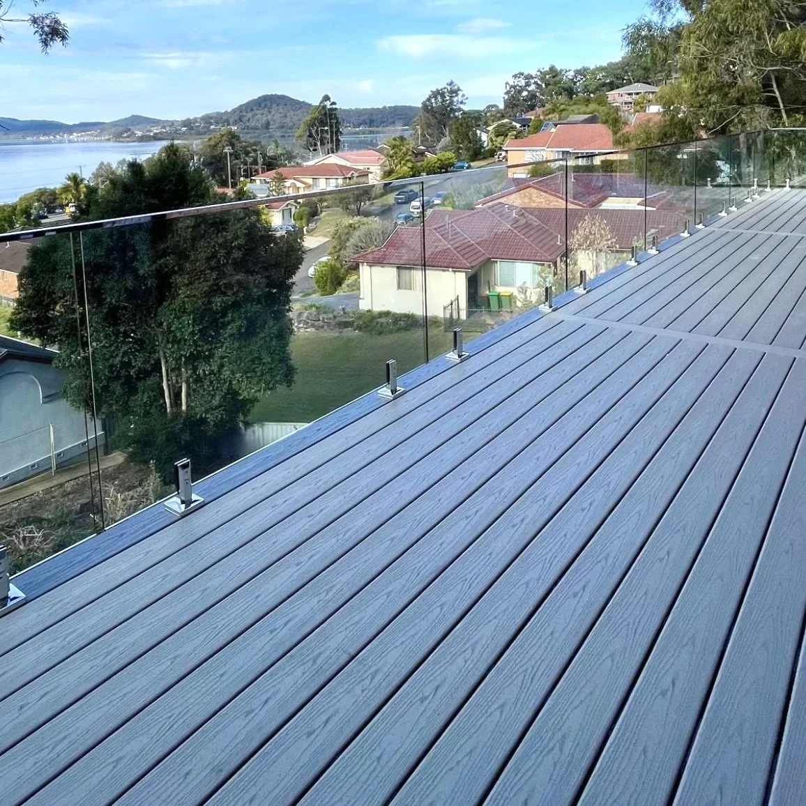 Elevated light grey composite deck with glass railing, overlooking a residential neighbourhood with houses, trees, and a hillside in the distance, with the ocean in the distance