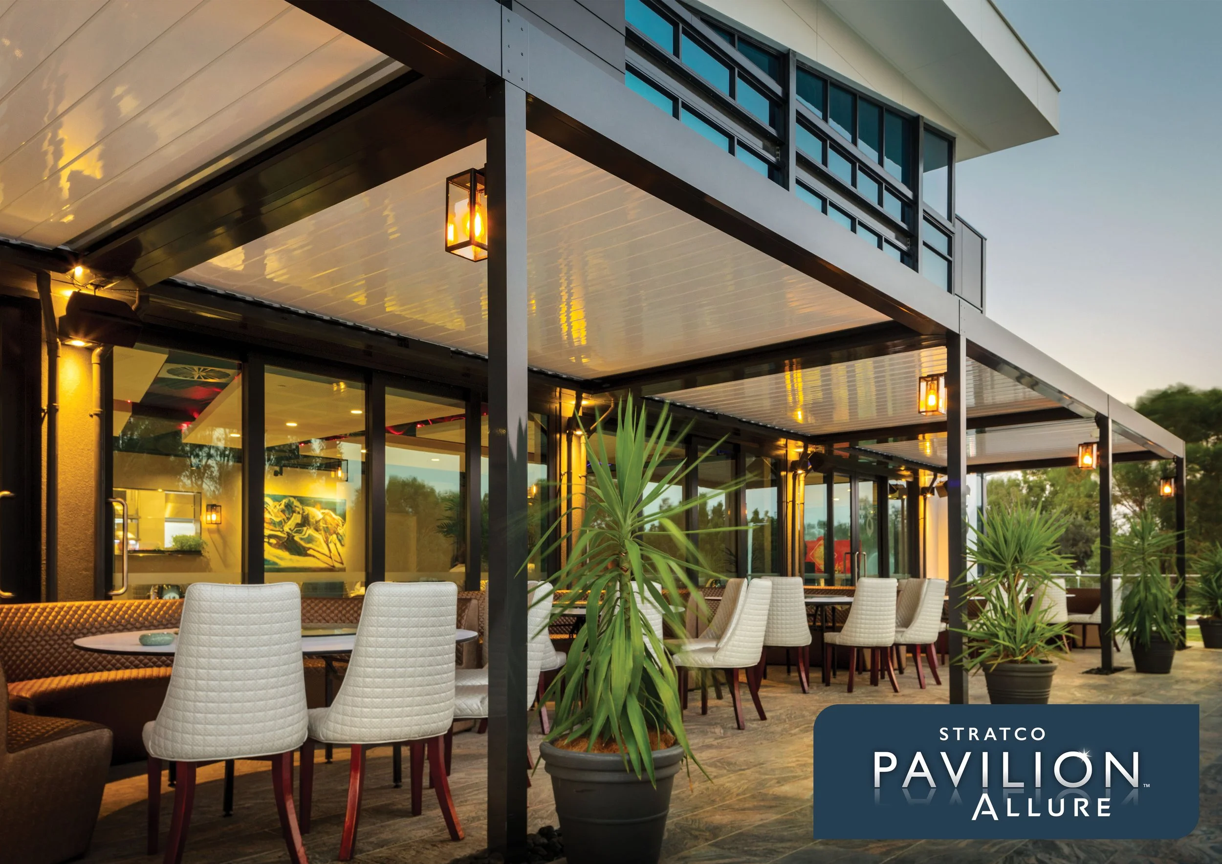 An outdoor  patio area of a modern building with tables and chairs, potted plants, and warm lighting fixtures, during dusk. Platinum Decks authorised installer.