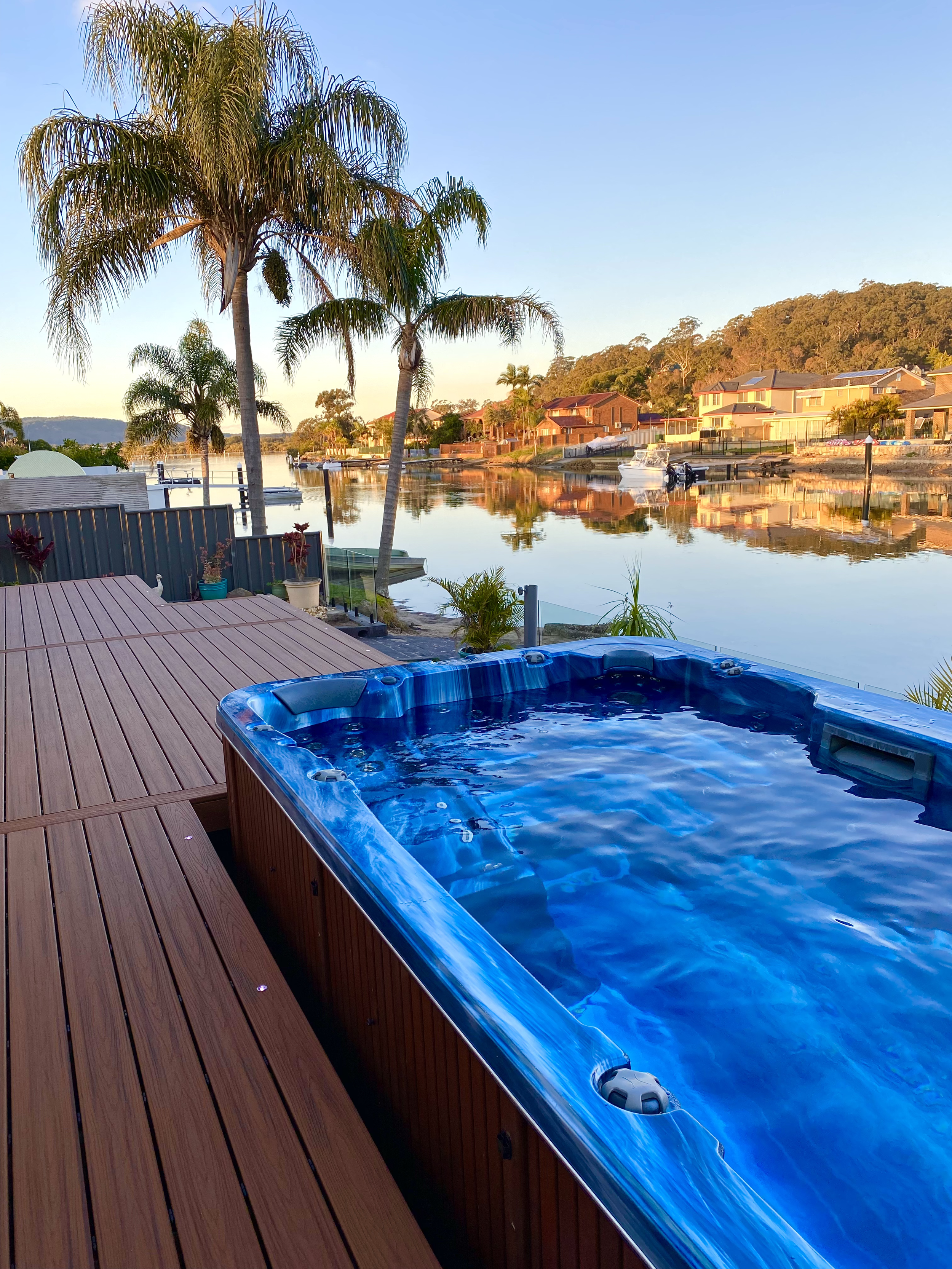 Platinum Decks built Composite deck built around waterfront spa overlooking a calm Brisbane Water channel at sunset with boats, palm trees and waterfront homes.