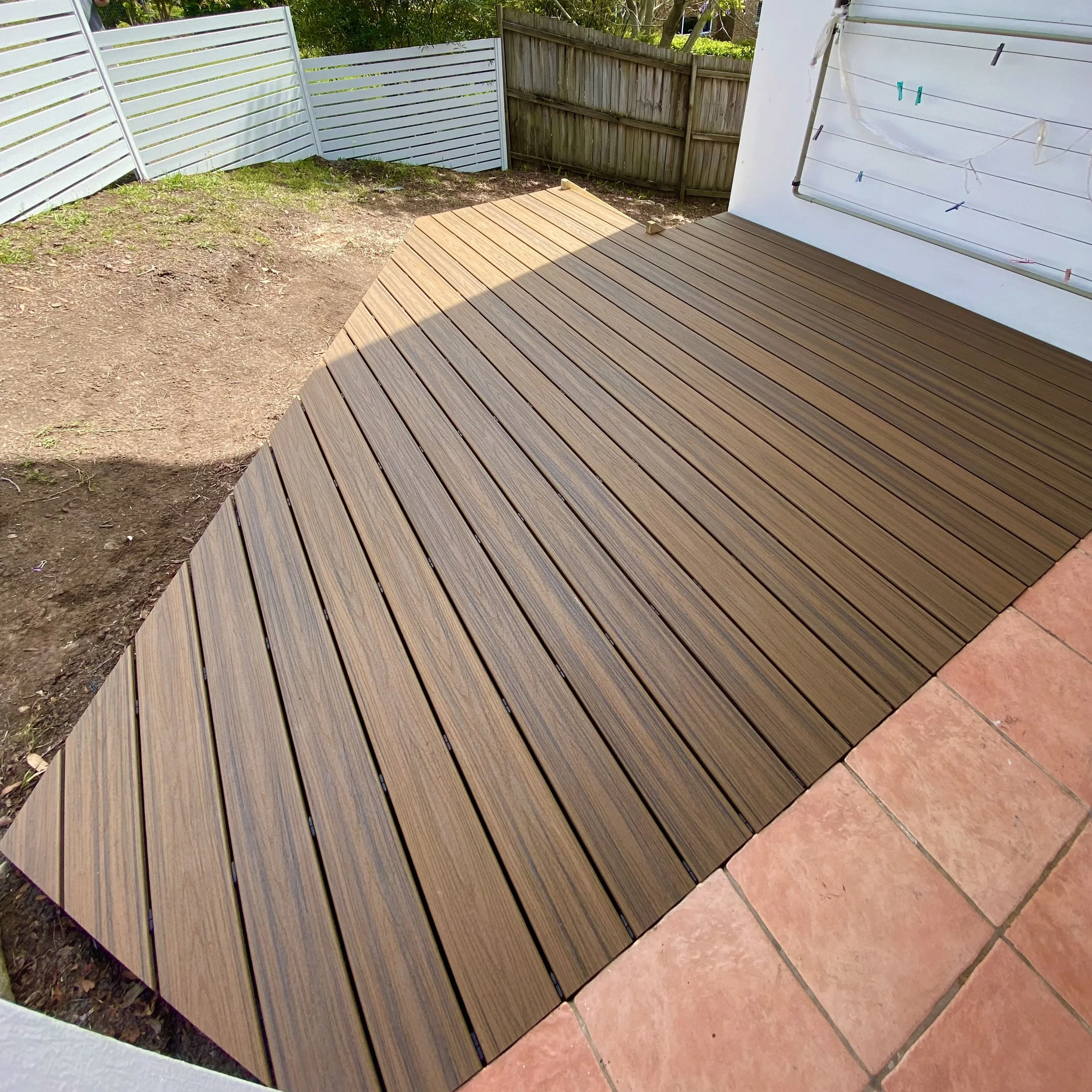 A backyard with a partially completed timber look composite deck, white fence, and a white wall with a clothesline and clothespins. Green foliage is visible in the background.