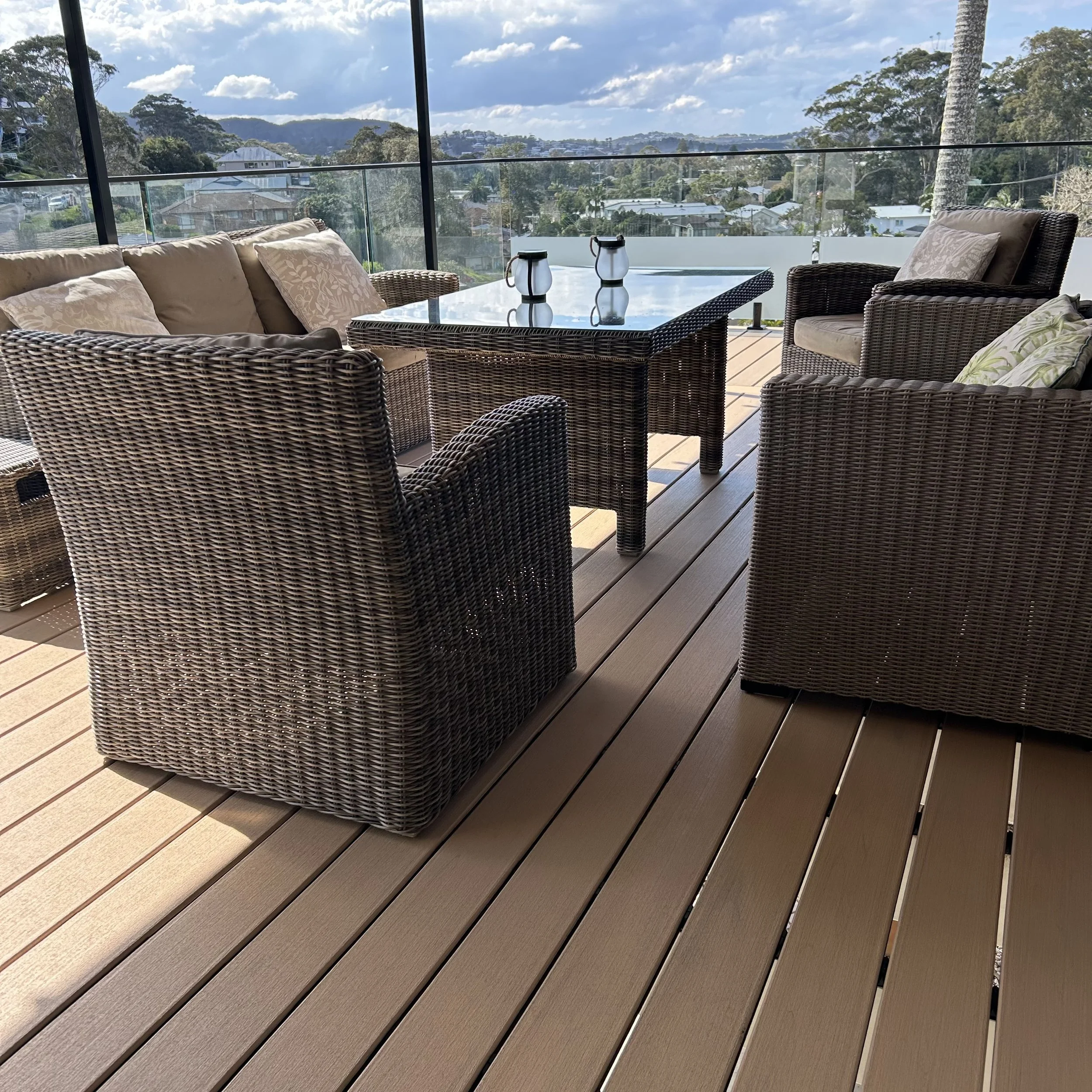 Outdoor composite deck and patio area with wicker furniture, glass table, and two lanterns, overlooking a scenic view of trees and hills under a partly cloudy sky on the Central Coast