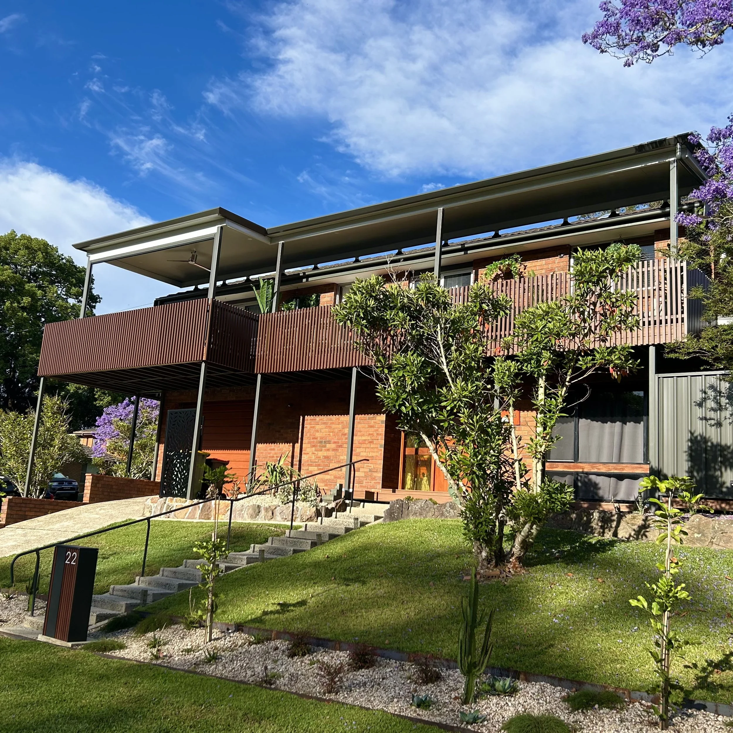 A modern two-story house with a brick exterior and large windows, surrounded by green lawn, trees, flowering plants, and a garden bed with rocks; blue sky with scattered clouds overhead.