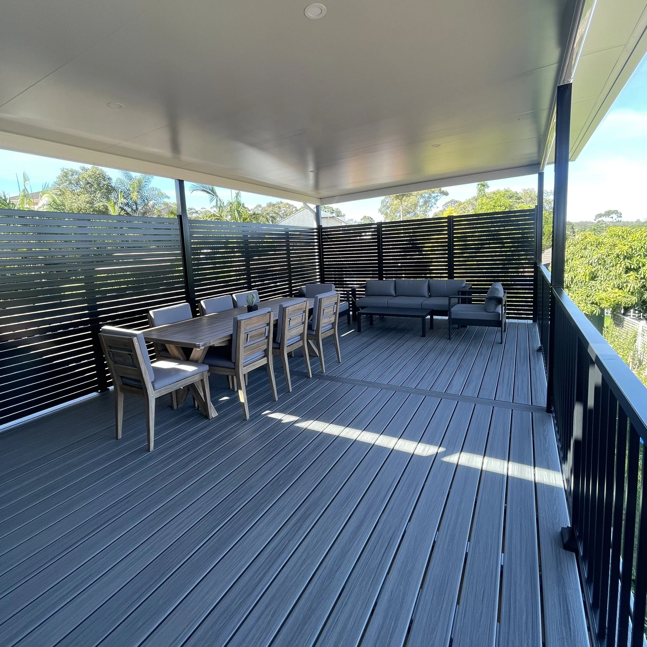 Backyard patio and composite deck with outdoor furniture, including a dining table, chairs, and a seating area with a sofa and coffee table, surrounded by a black horizontal slat fence, with trees and a partly cloudy sky in the background.