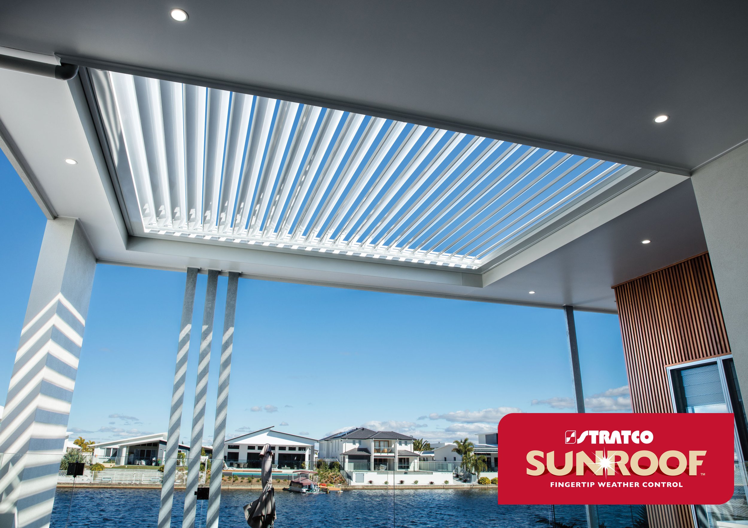View of a waterfront neighborhood with modern houses, swimming pool, and palm trees under a clear blue sky, seen from a covered patio with solar sunroof panels. Platinum Decks authorised Outback Dealer.