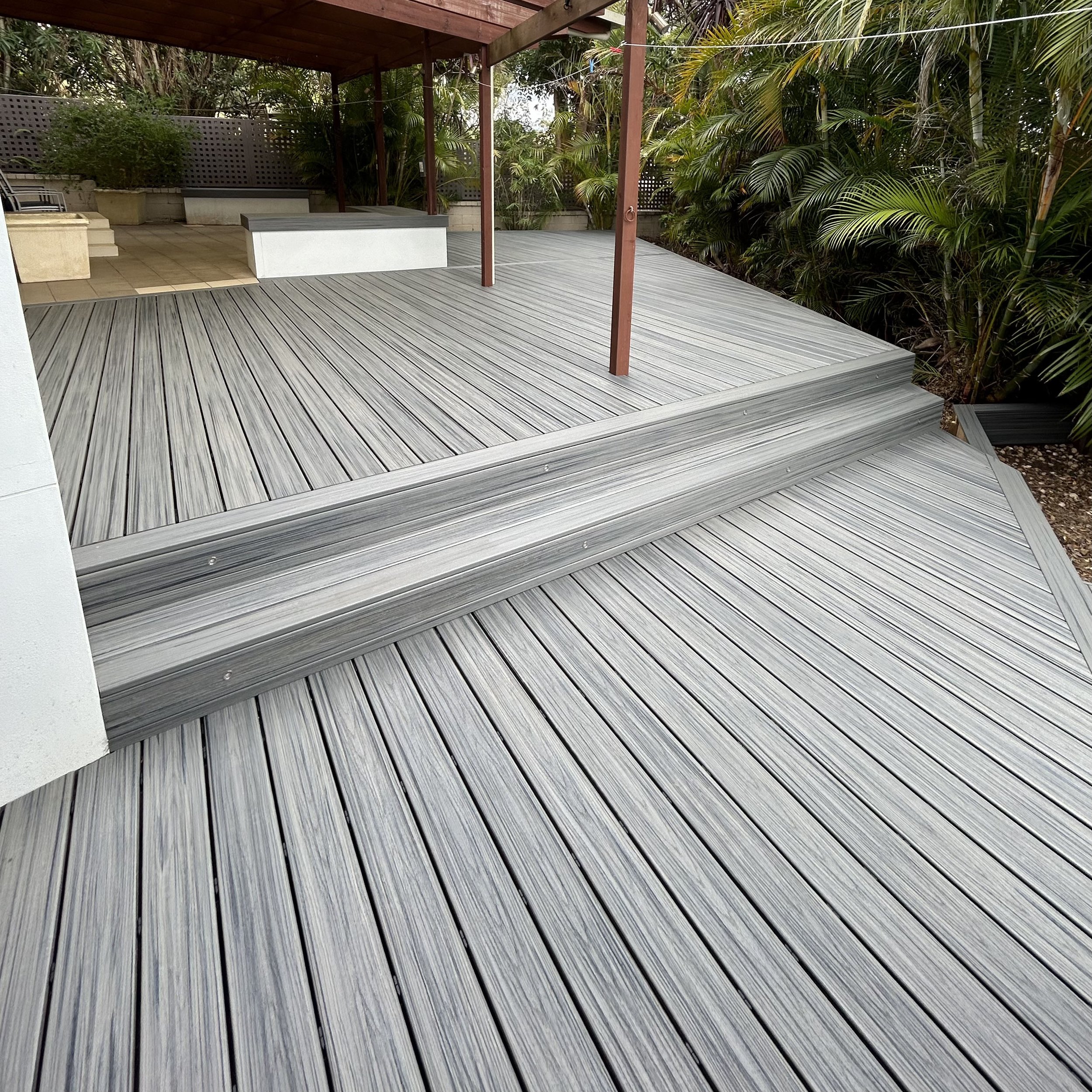 A wooden deck with horizontal planks, stairs leading down, a covered area with a roof, and lush green tropical plants surrounding the space.