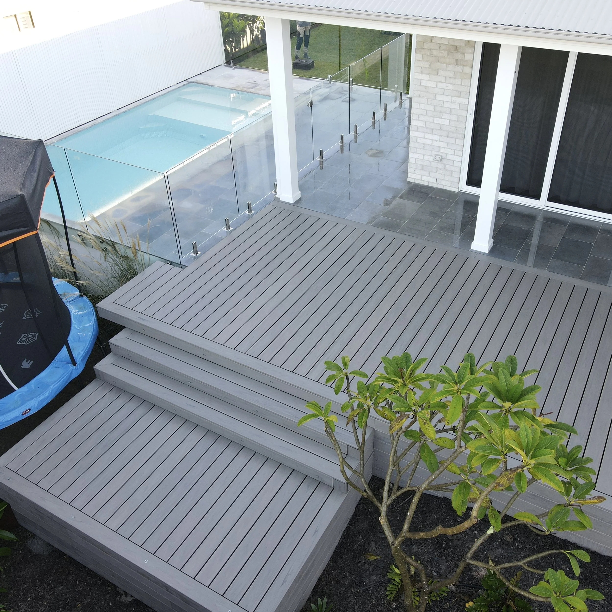 A backyard patio area with a small pool, glass fencing, tree, and sliding glass doors.