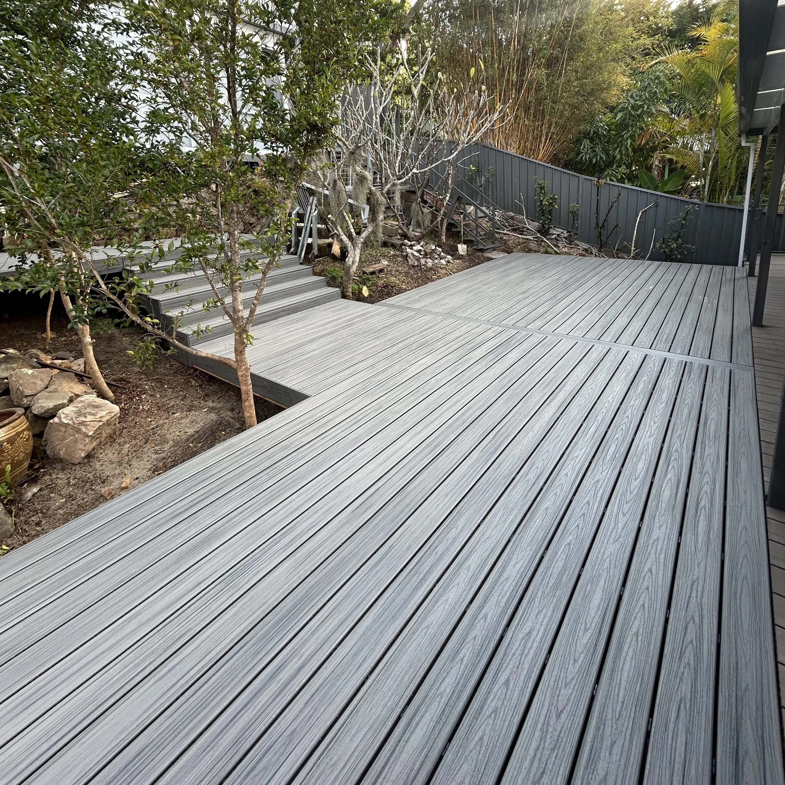 A modern composite deck extends from a patio into a backyard with trees and bushes, enclosed by a fence, with some greenery and a metal staircase in the background.