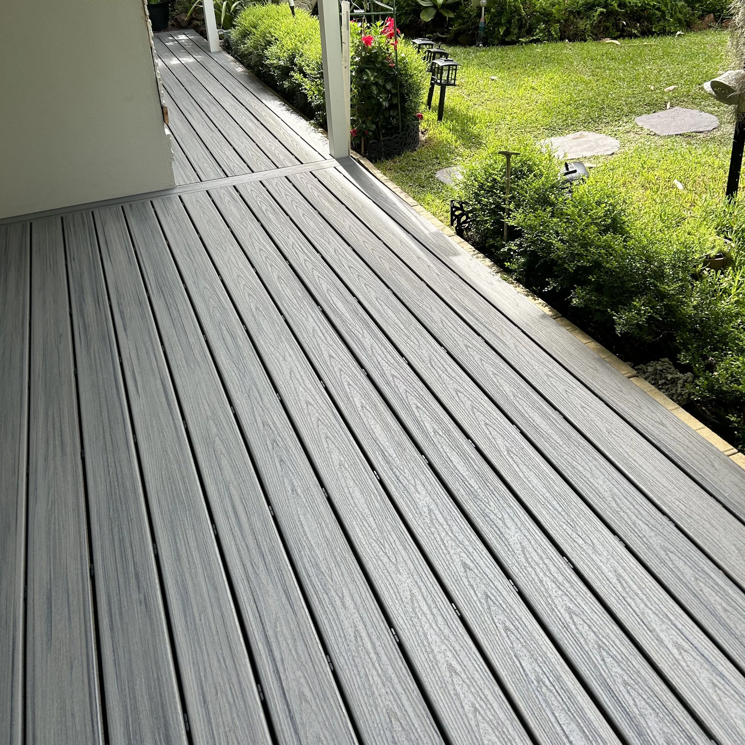 An outdoor wooden deck next to a garden with green bushes, pink flowers, stone pathway, and garden lights.