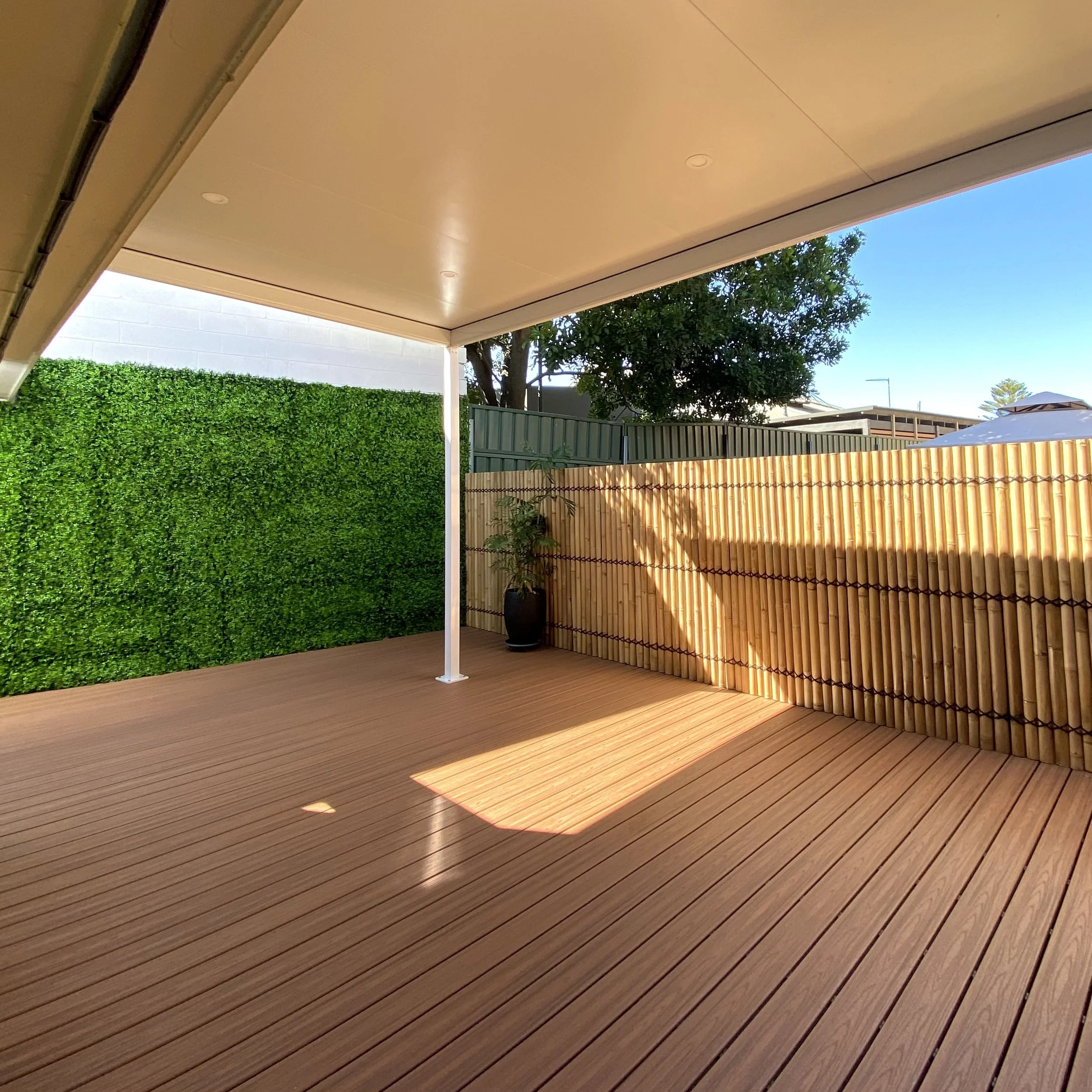 Stunning backyard composite deck and patio area with a tall privacy fence, a green hedgerow, a potted plant, and custom patio overhead. Clear blue sky and trees are visible in the background.