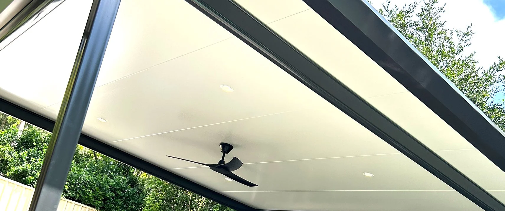 Close-up of a modern, covered outdoor patio with a ceiling fan and recessed lighting, surrounded by greenery and trees.