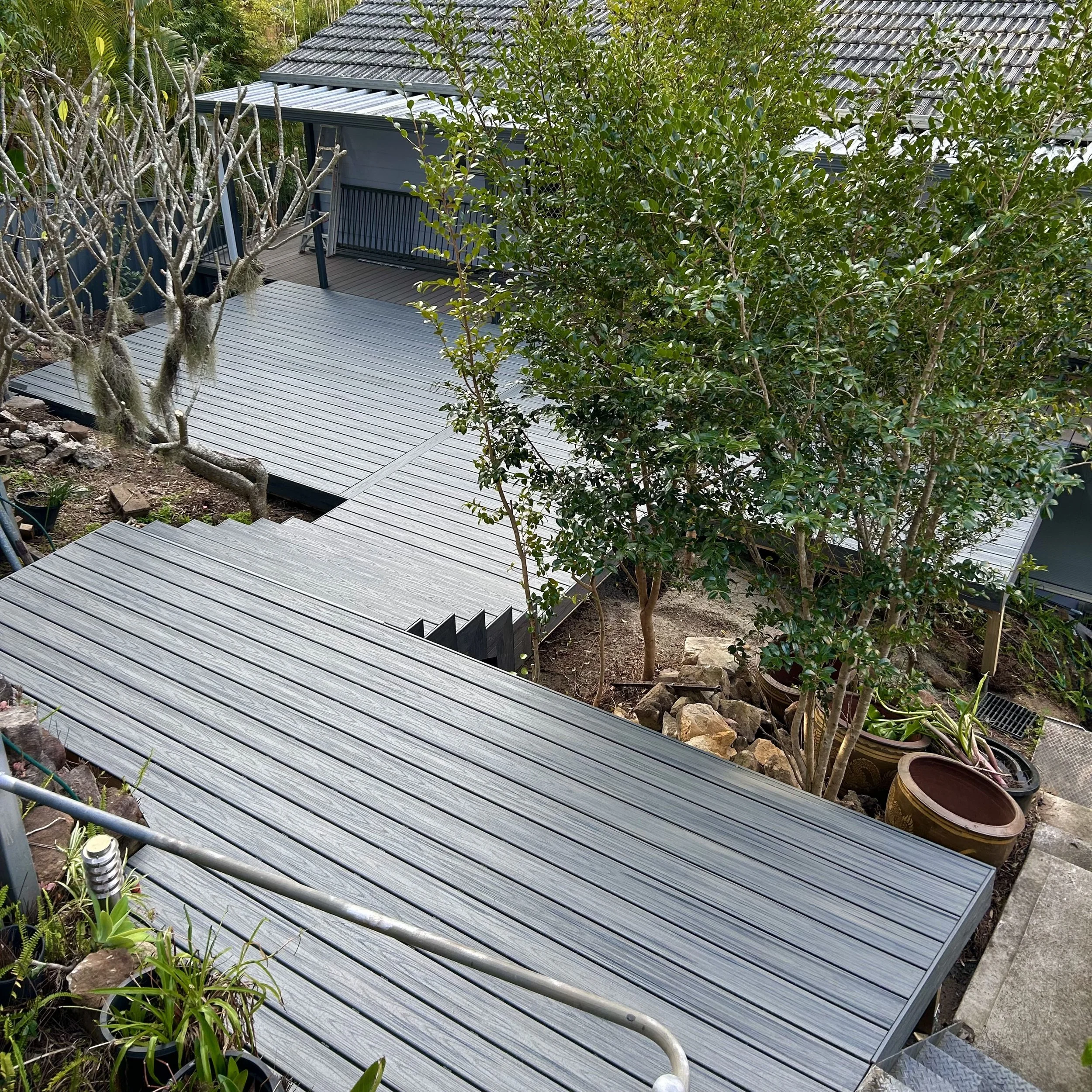 A backyard oasis architecturally designed by Plainum Decks consisting of a large composite deck surrounded by trees and plants, including a large leafy bush and a bare tree with hanging moss.