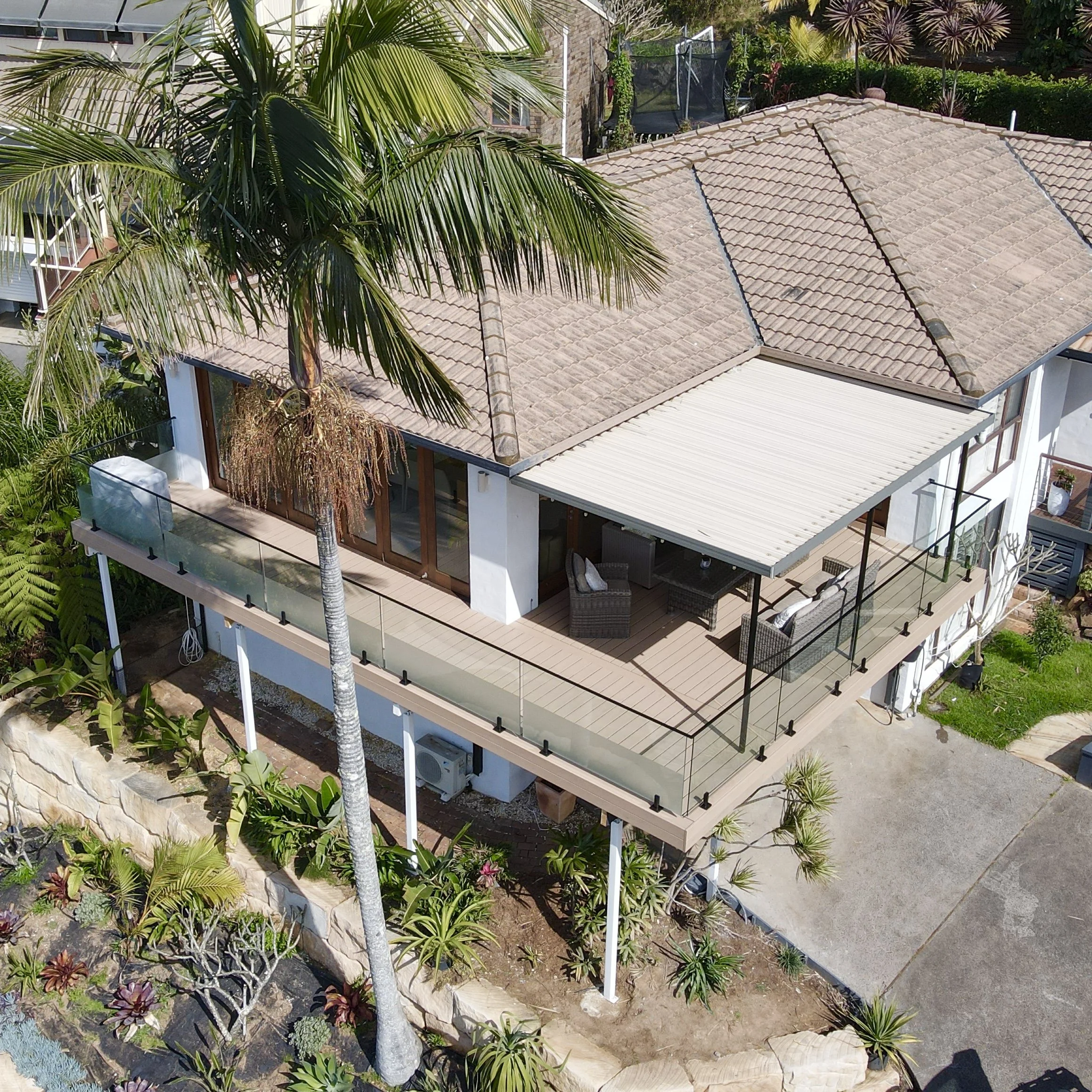 Two-story house with beige tiled roof, surrounded by green plants and trees, including a tall palm tree, with a balcony and outdoor seating area.