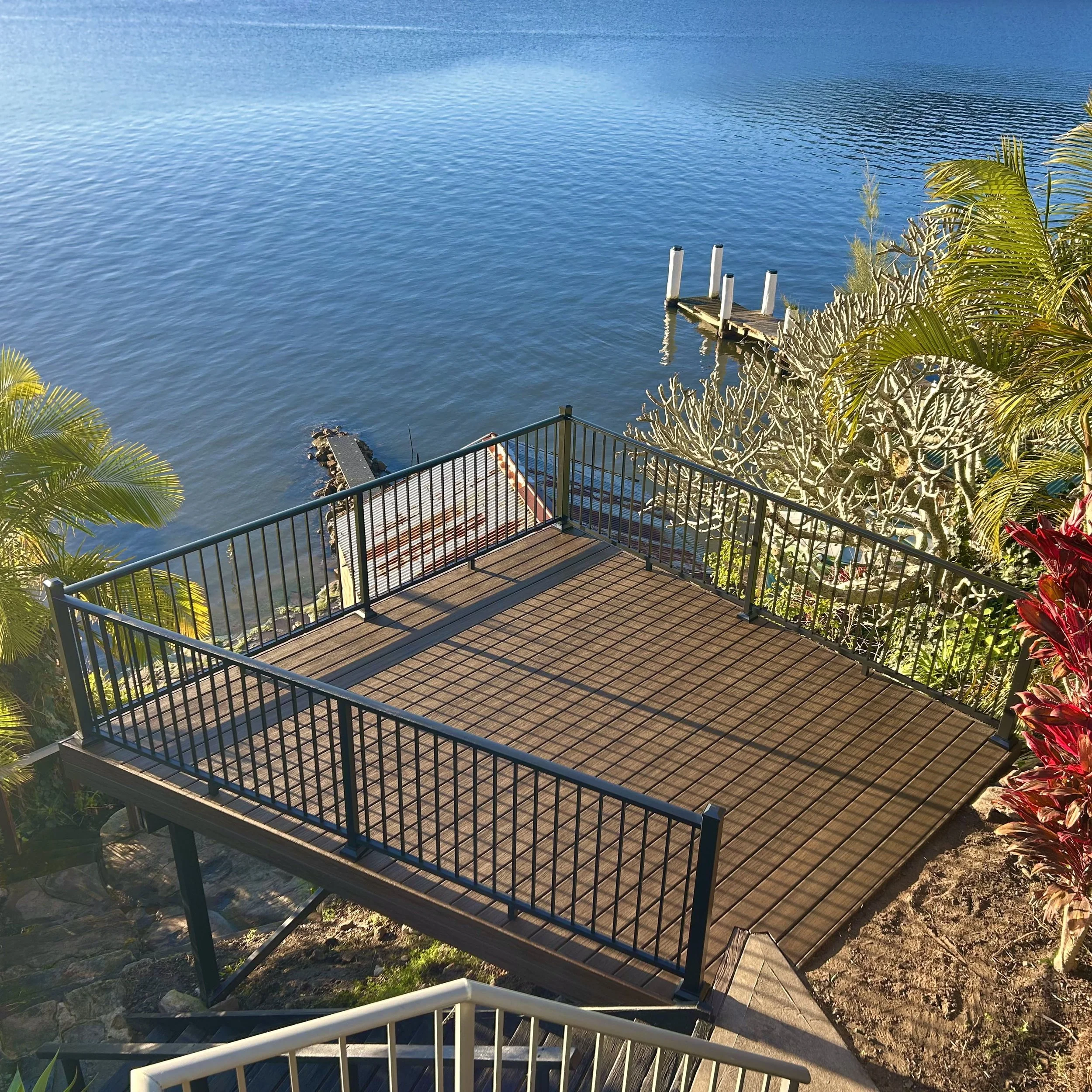 View of a blue lake with a Trex composite deck and railing along the shoreline, surrounded by palm and other tropical plants.