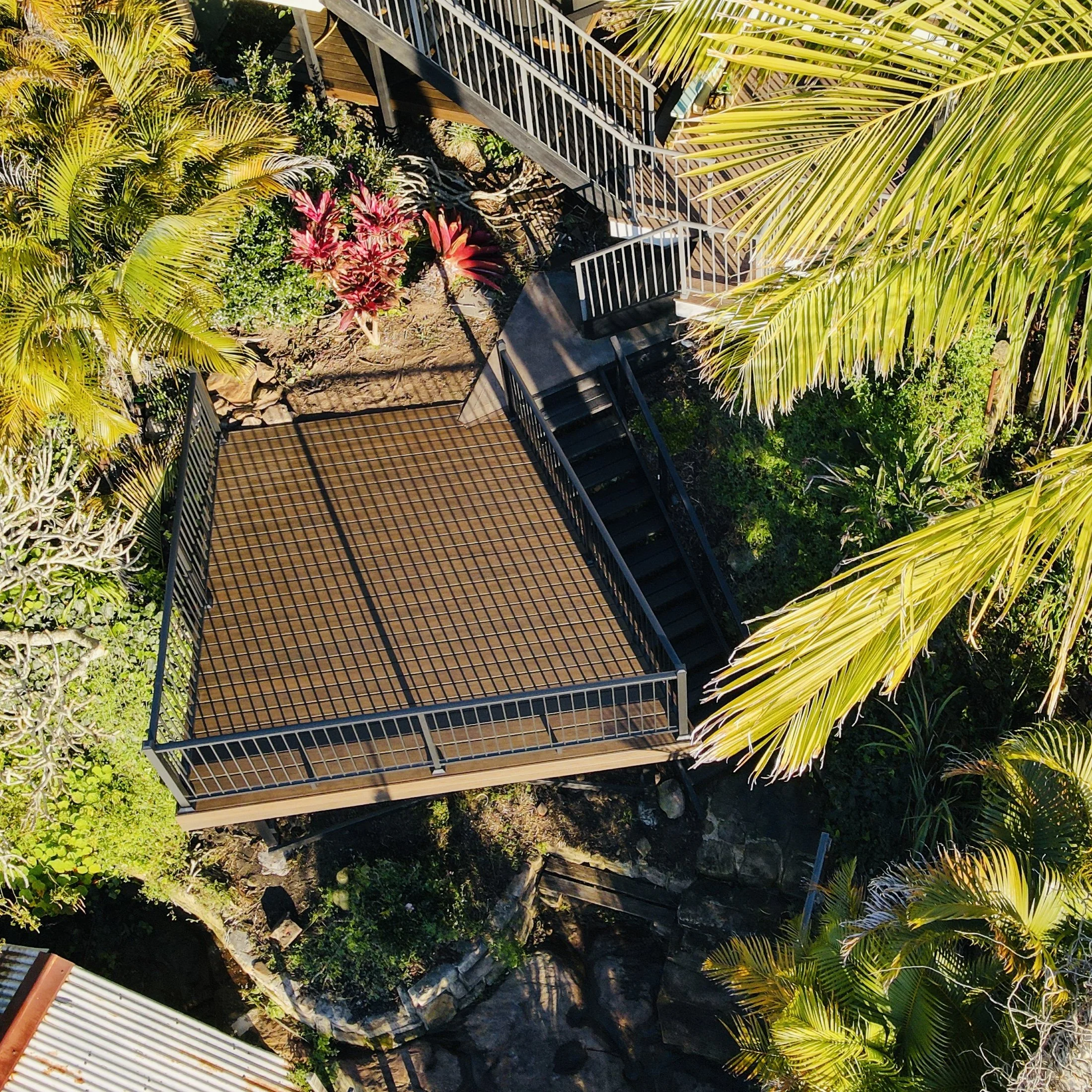Top-down view of a composite deck surrounded by tropical plants, including palm trees and vibrant pink flowering bushes, with a white metal railing and stairs leading down to a lower area.
