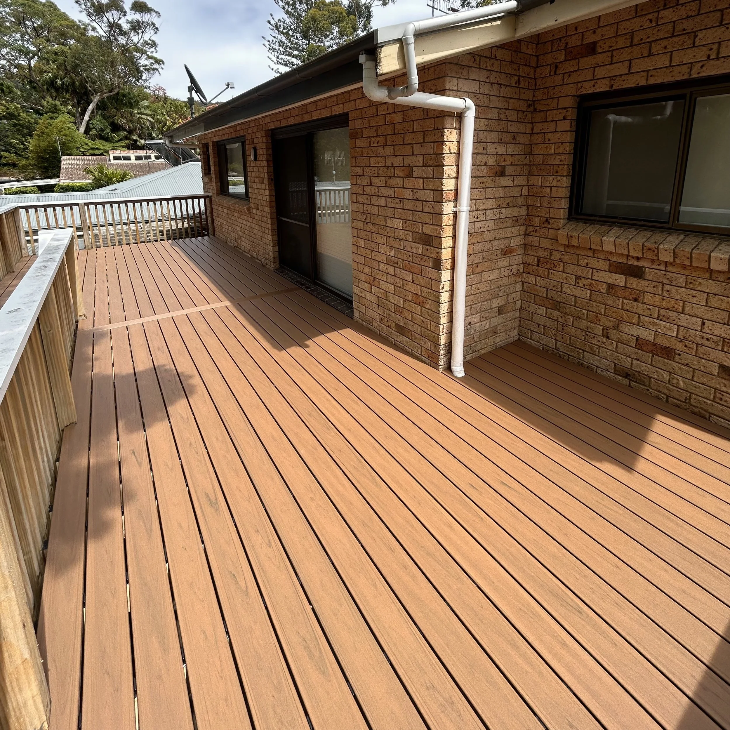 A large fresh and modern outdoor composite deck attached to a brick house, with a railing on the left side. The house has sliding glass doors and a window, with white gutter and downspout.
