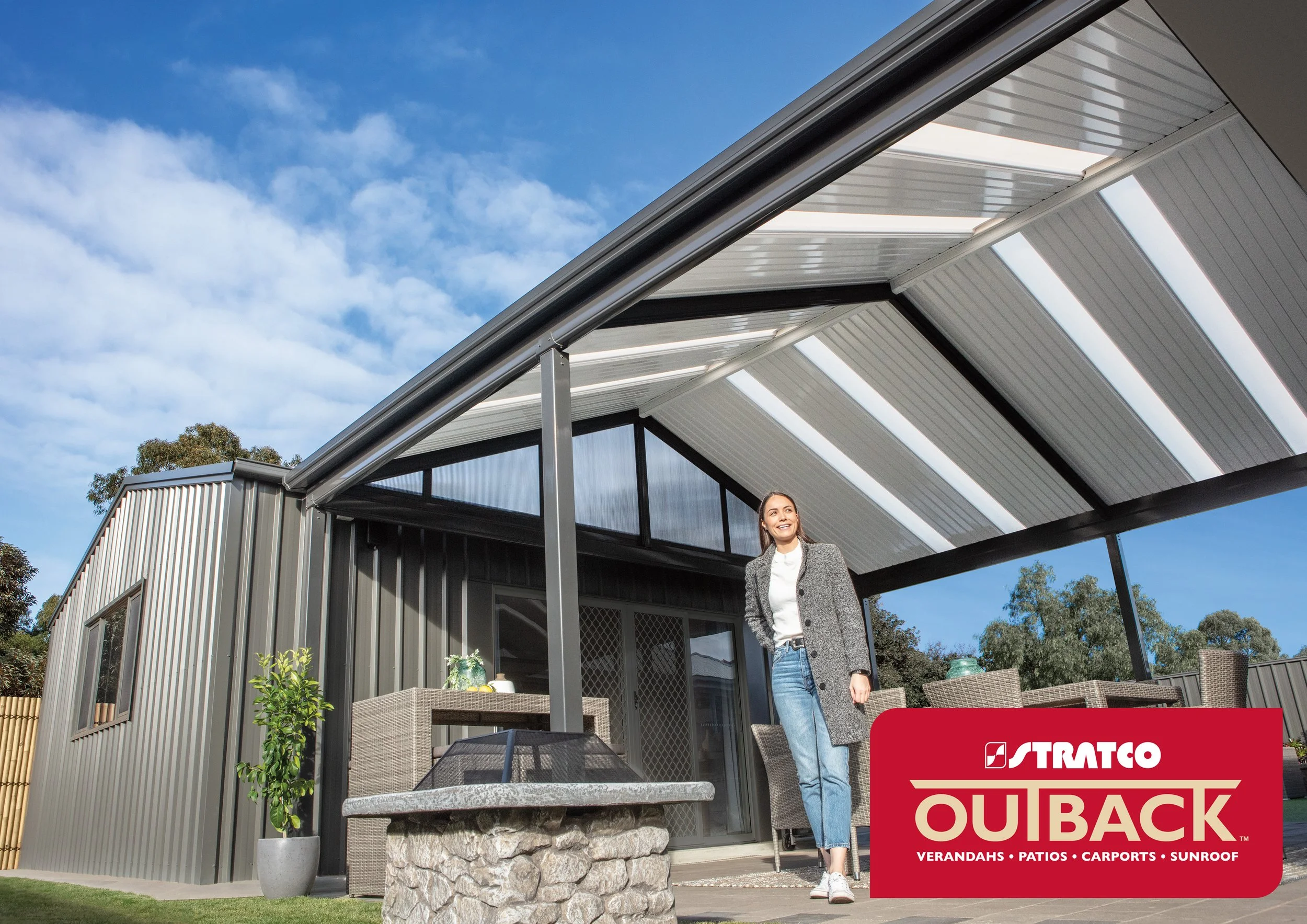 A woman standing outside a modern house with a patio and sunshade. The house has gray siding and large windows. There is a table and chairs on the patio. A red logo with white and gold text reads "Stratco Outback, Verandahs, Patios, Carports, Sunroom