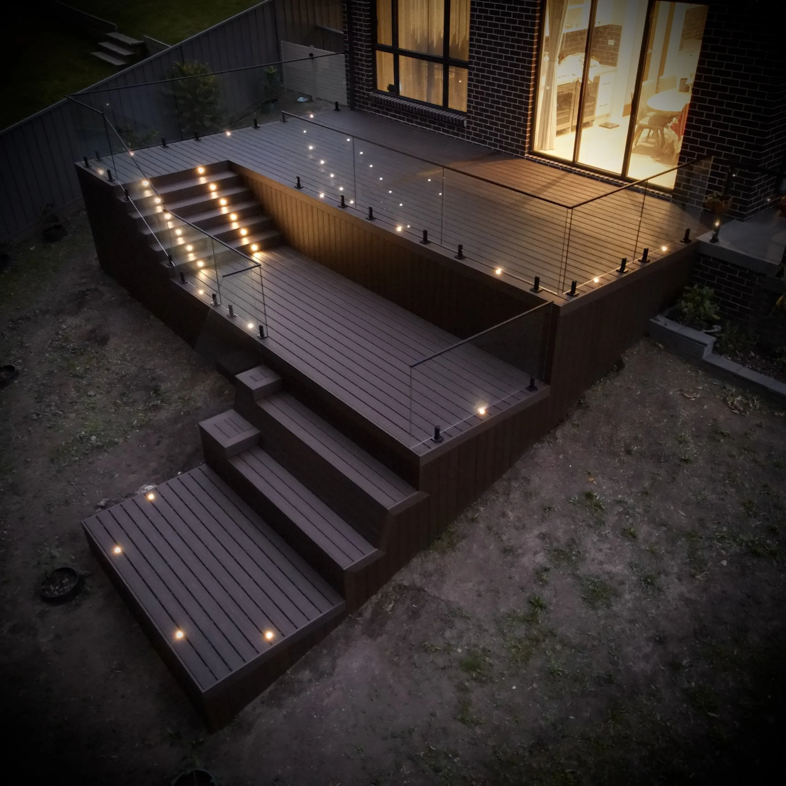 A nightime view of a Tiered Trex composite deck with integrated lighting and steps by Platinum Decks & Patios for modern entertaining