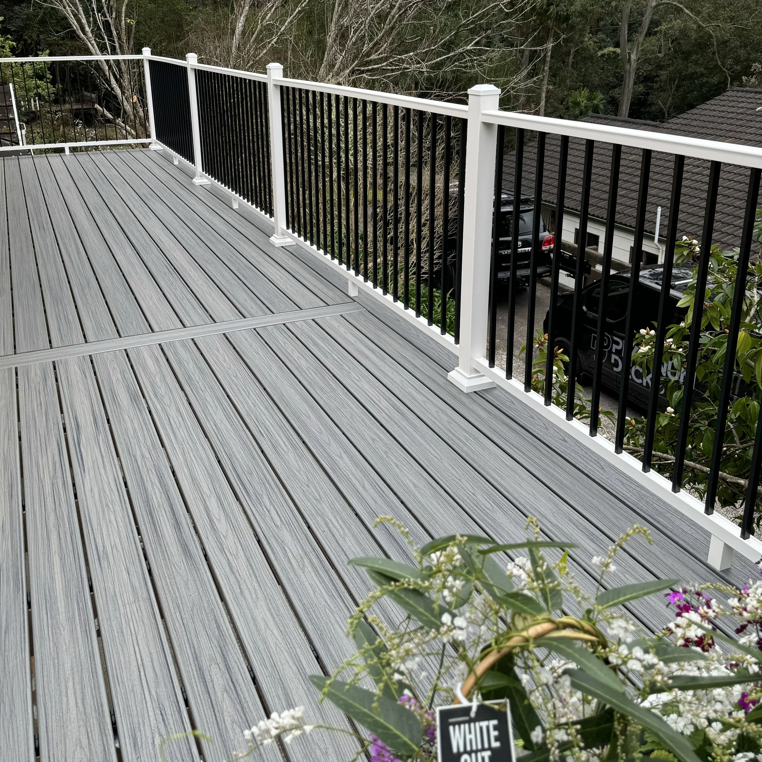 A modern grey composite Trex deck and black and white metal railings, overlooking a yard with trees and a house with a tiled roof, some cars parked, and colourful flowers in the foreground.