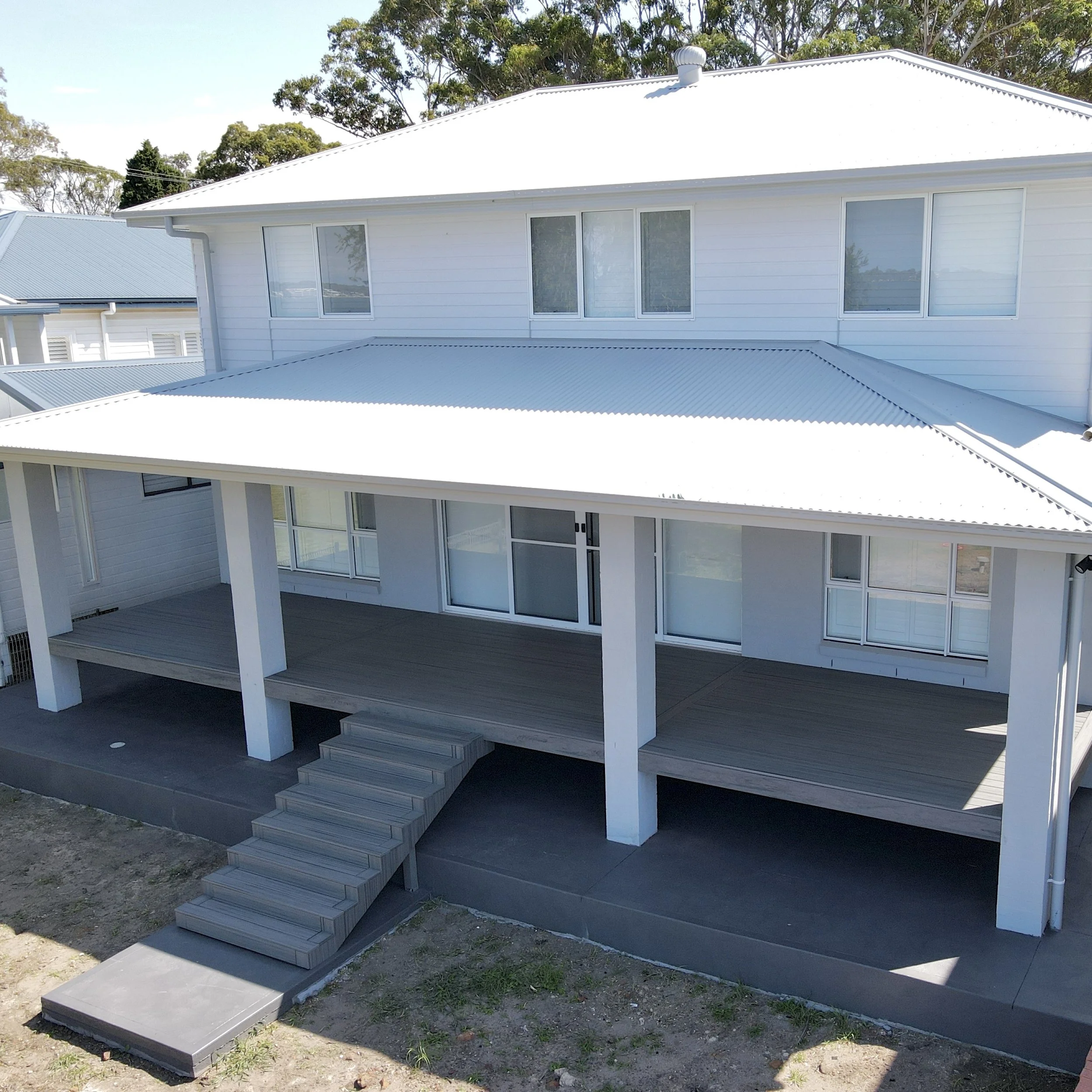 White modern two-story house with metal roof, large windows, a small staircase leading to a deck, and multiple sections with similar roofing. Trees in the background, grey paved pathway in front.