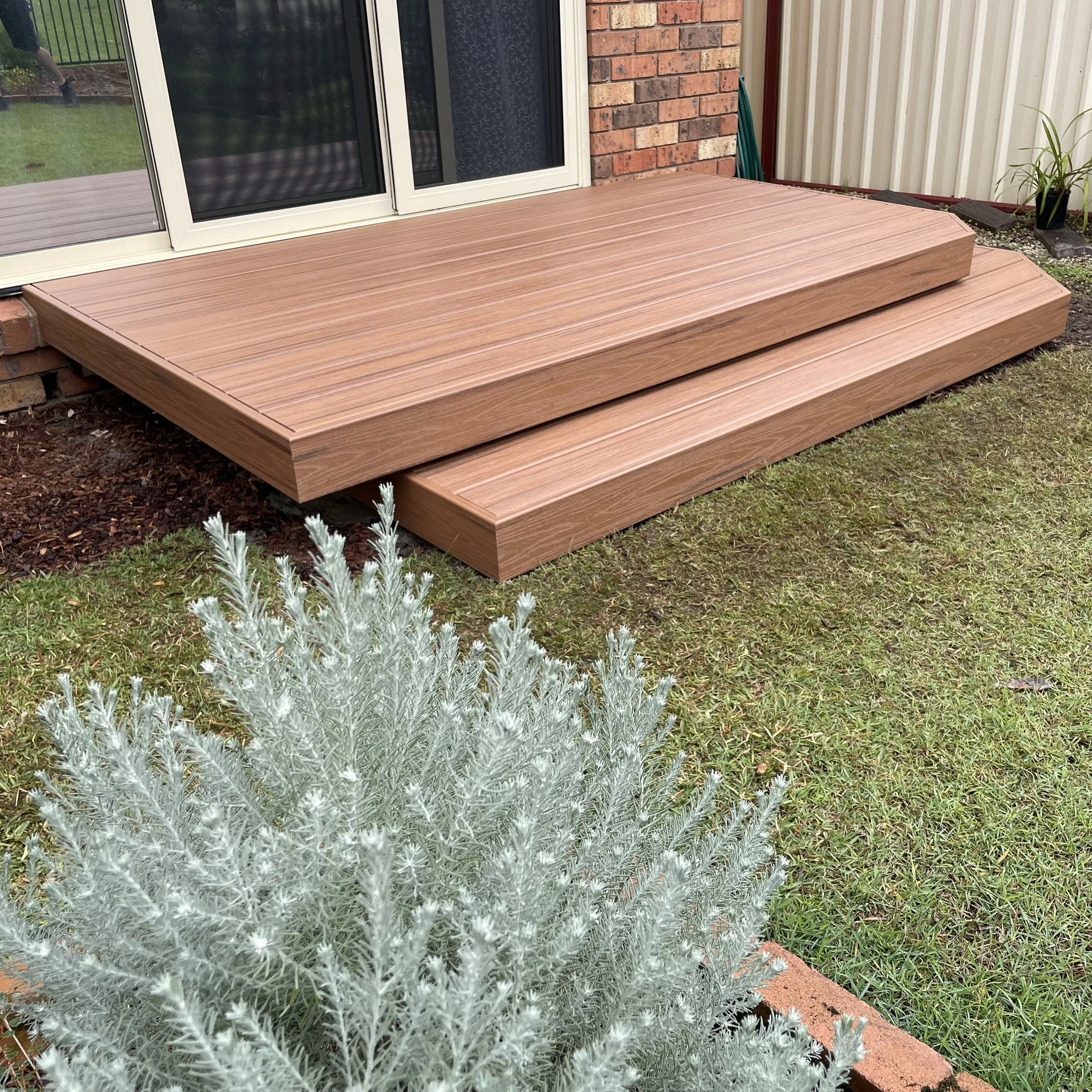 Trex composite deck in tiki torch beside brick home and garden elements installed by Platinum Decks to blend high‑end craftsmanship with natural outdoor textures