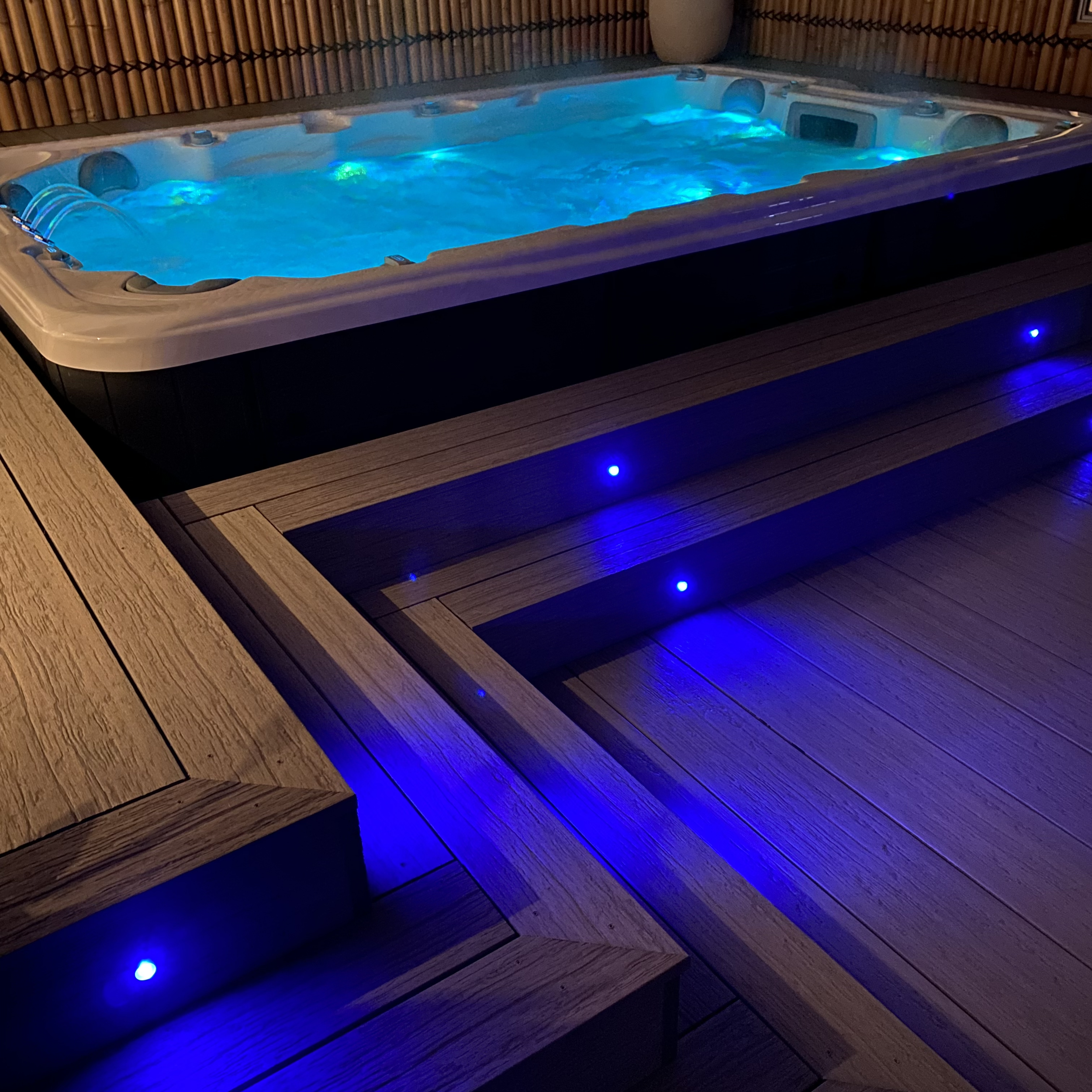 Outdoor spa glowing at night with blue underwater lights, surrounded by rich brown composite deck and stairs and a tall bamboo privacy fence built and designed by Platinum Decks.