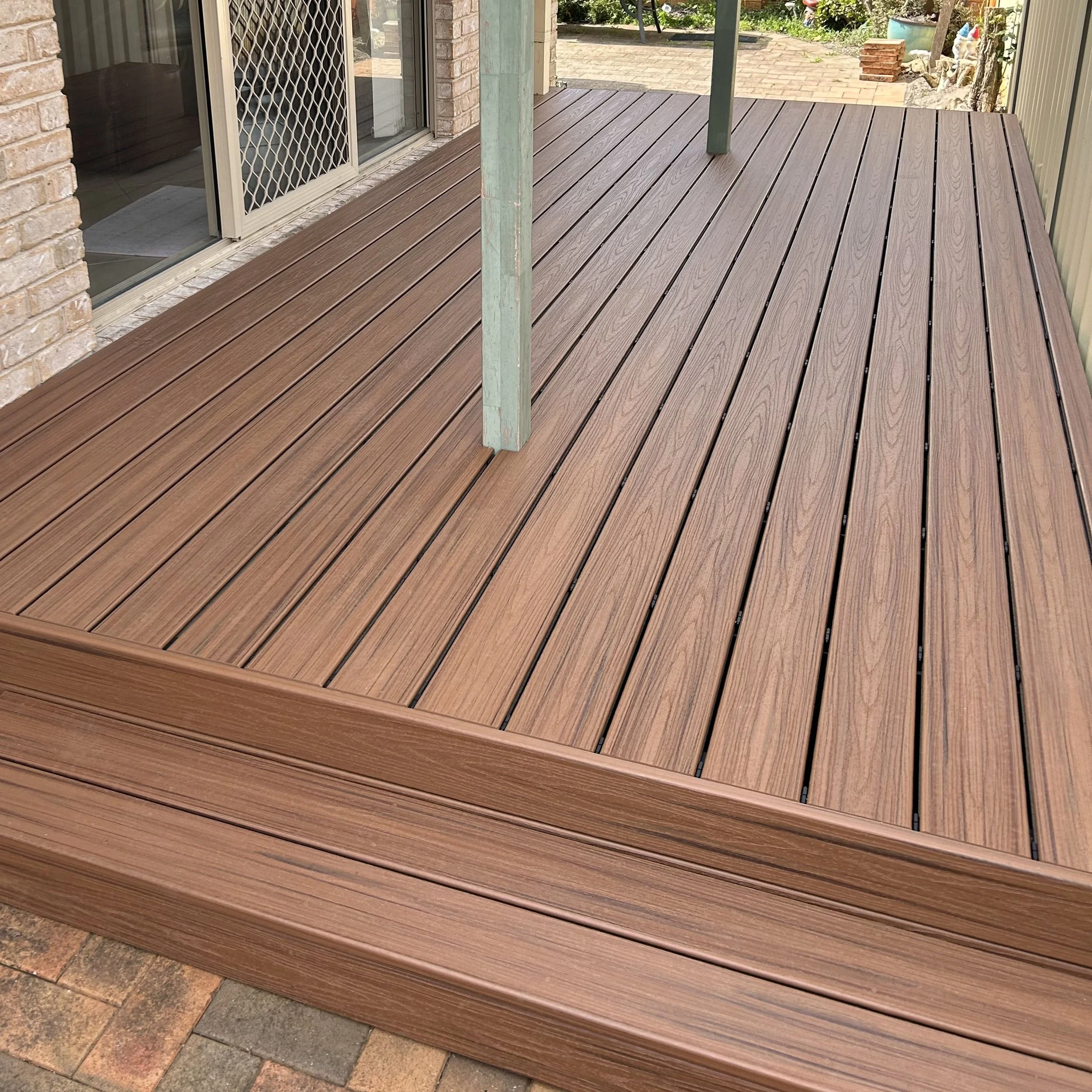 A modern fresh composite deck attached to a suburban Central Coast house, with sliding glass doors and a metal support pole, overlooking a backyard with garden decorations and a paved area.