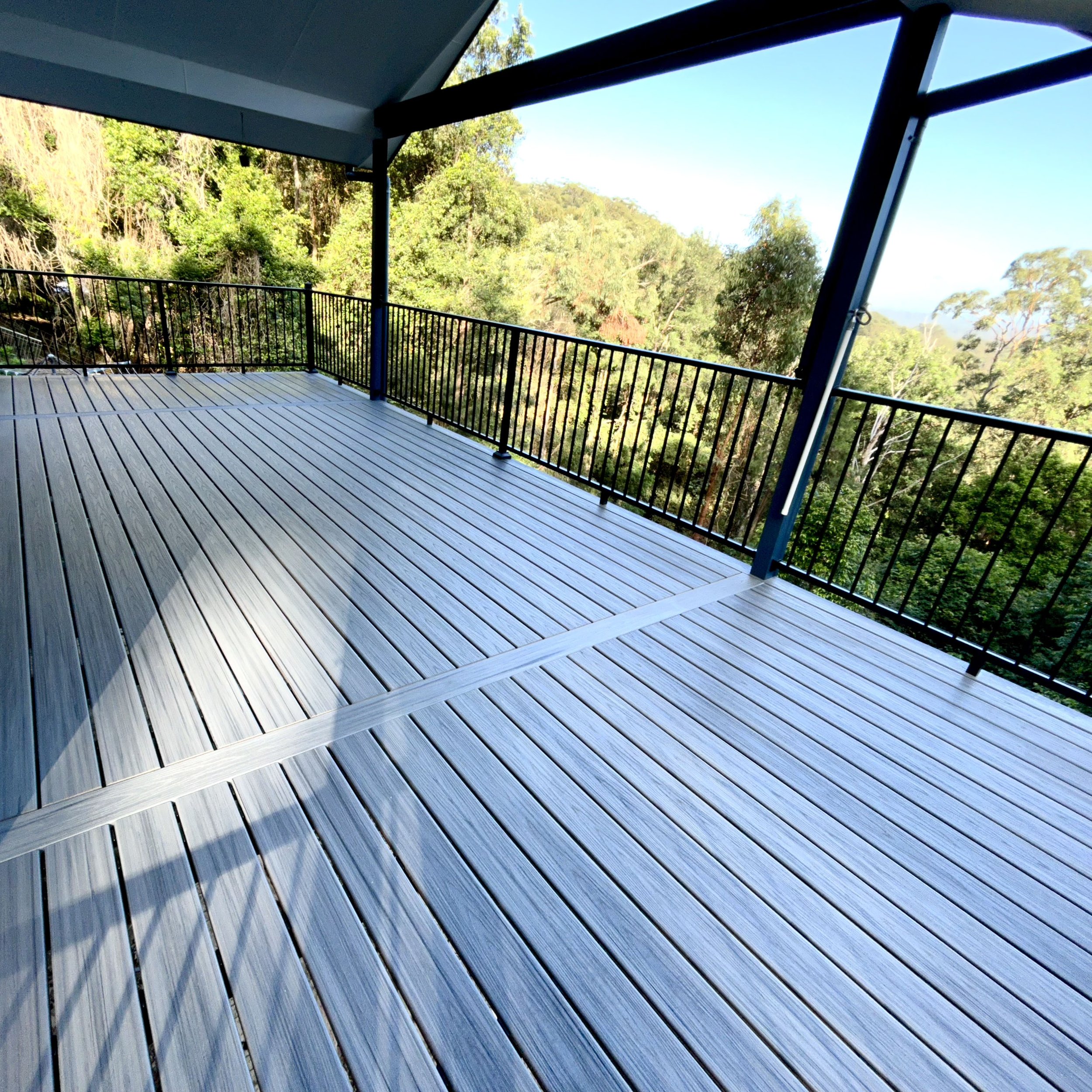 View from a deck showing a newly installed composite deck with a black metal railing and a backdrop of lush green trees under a clear blue sky.