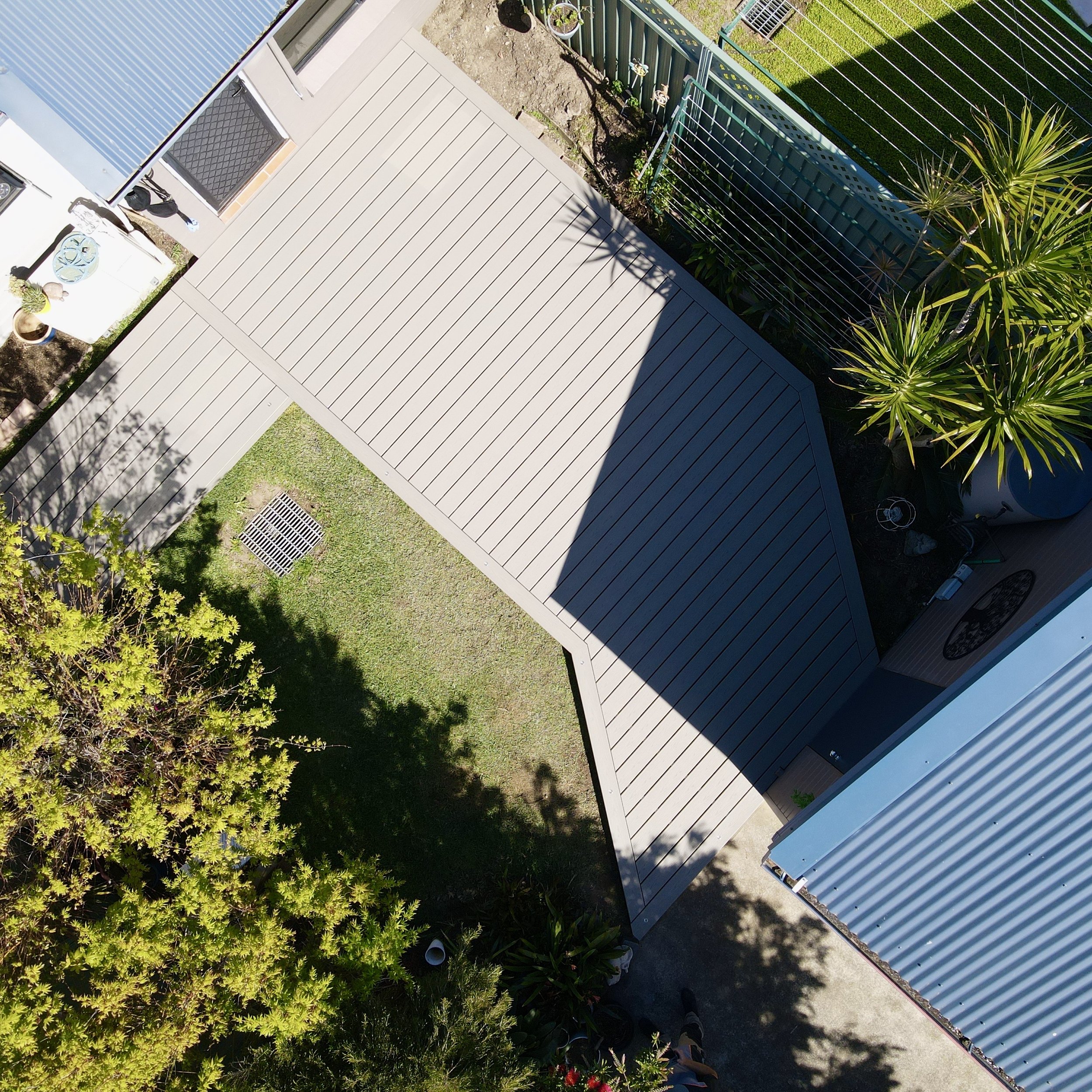 An aerial view of a backyard with a wooden deck, a garden bed, a green metal fence, and various plants including trees and shrubs.