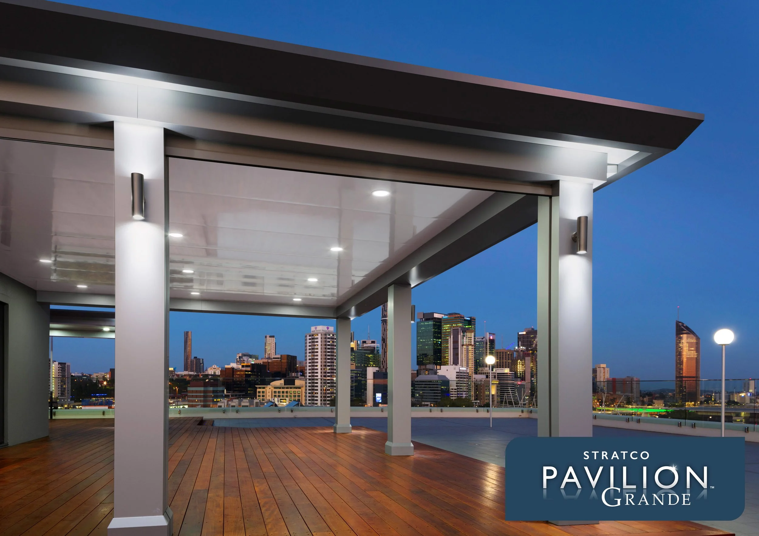 View of a modern patio roof with a wooden deck, white pillars, and a city skyline in the background during twilight. The pavilion has a sleek roof and is illuminated by integrated lights. Platinum Decks authorised installer.