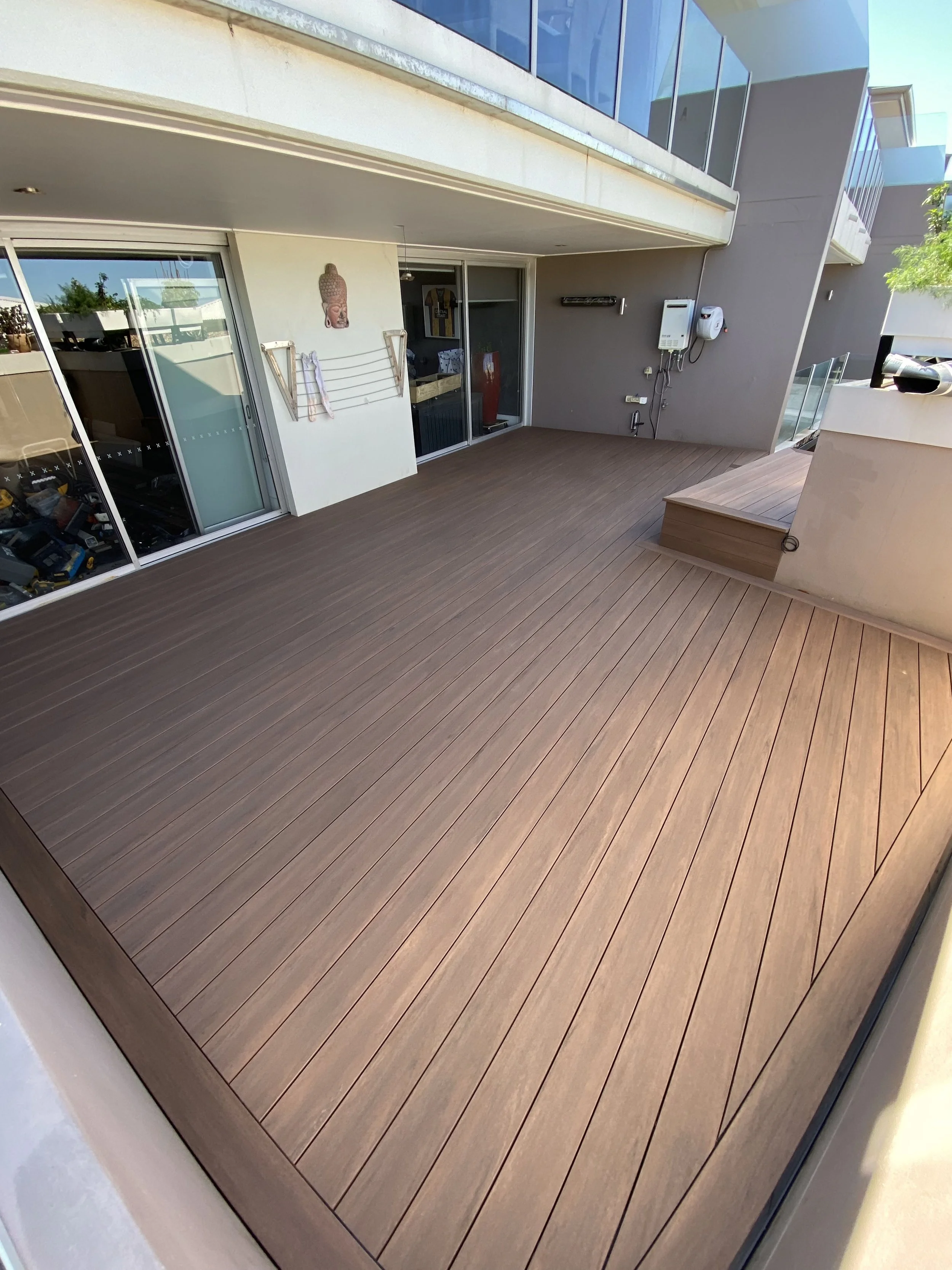 A modern outdoor balcony with wooden flooring, glass sliding doors, and a decorative Buddha wall hanging, with some objects inside the apartment visible through the glass.