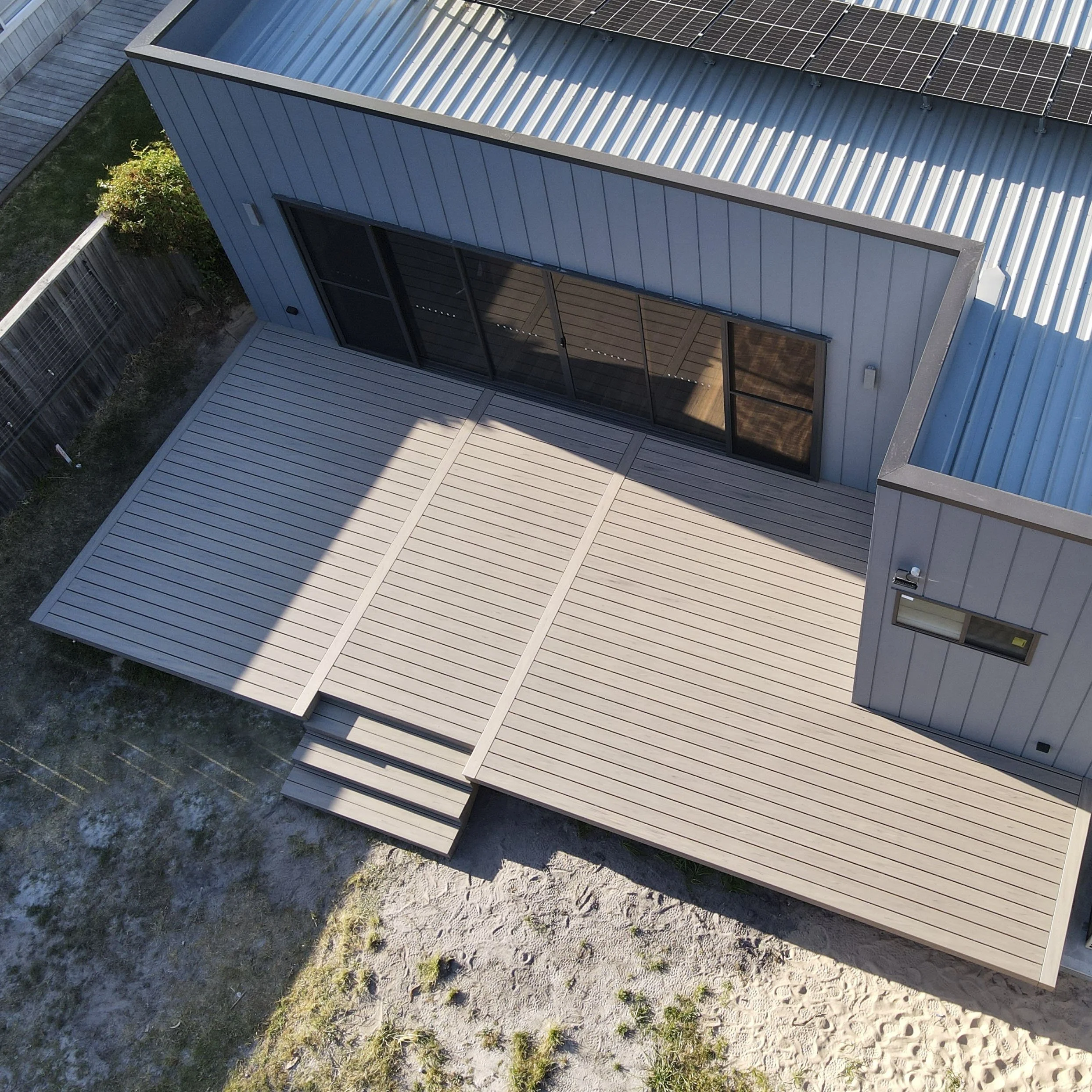 A modern house with a metal roof featuring solar panels, glass sliding doors, a wooden deck, and a small garden area with grass and stones.