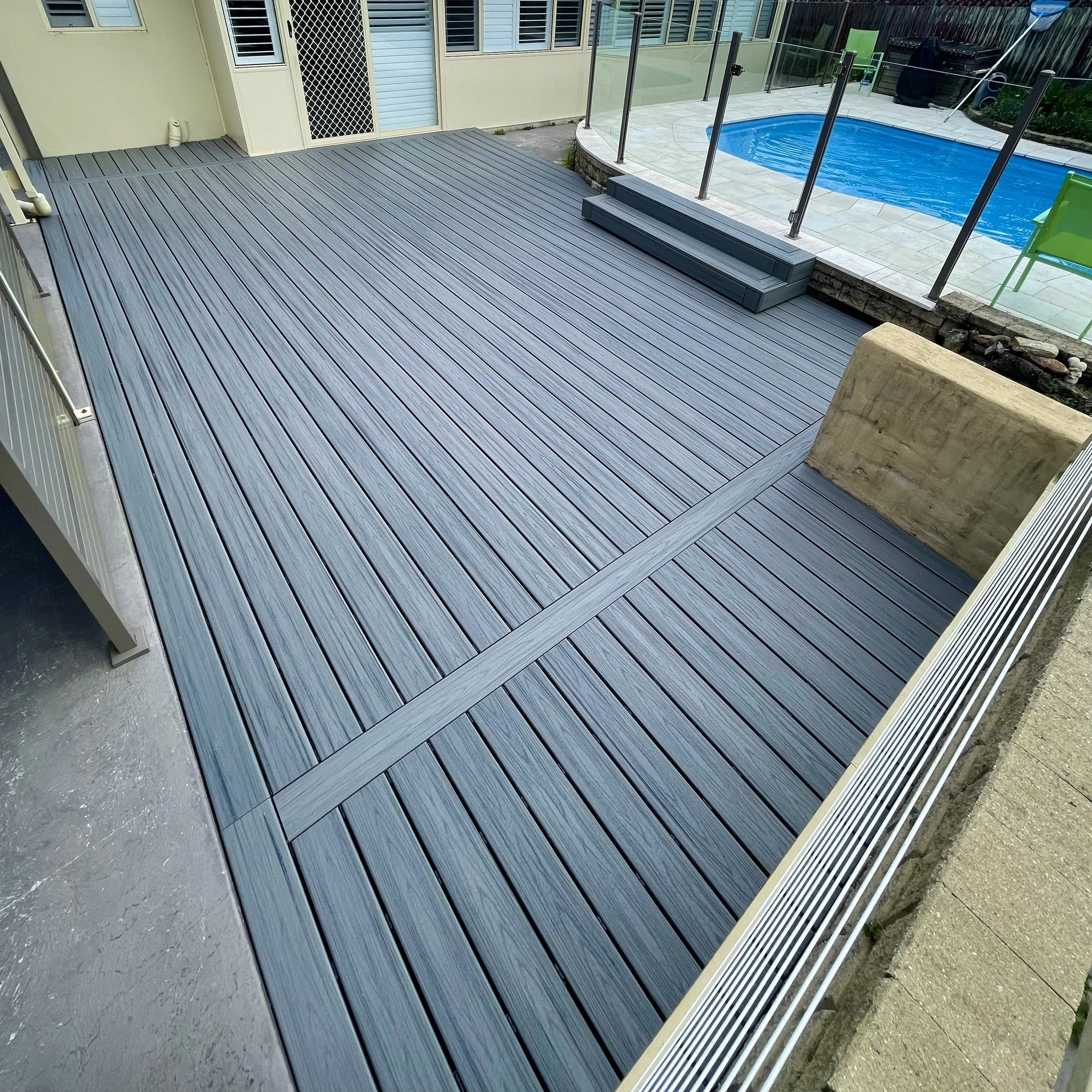 A backyard patio with gray wood decking, a small set of steps, and a glass fence surrounding a blue swimming pool, with some outdoor furniture and plants in the background.