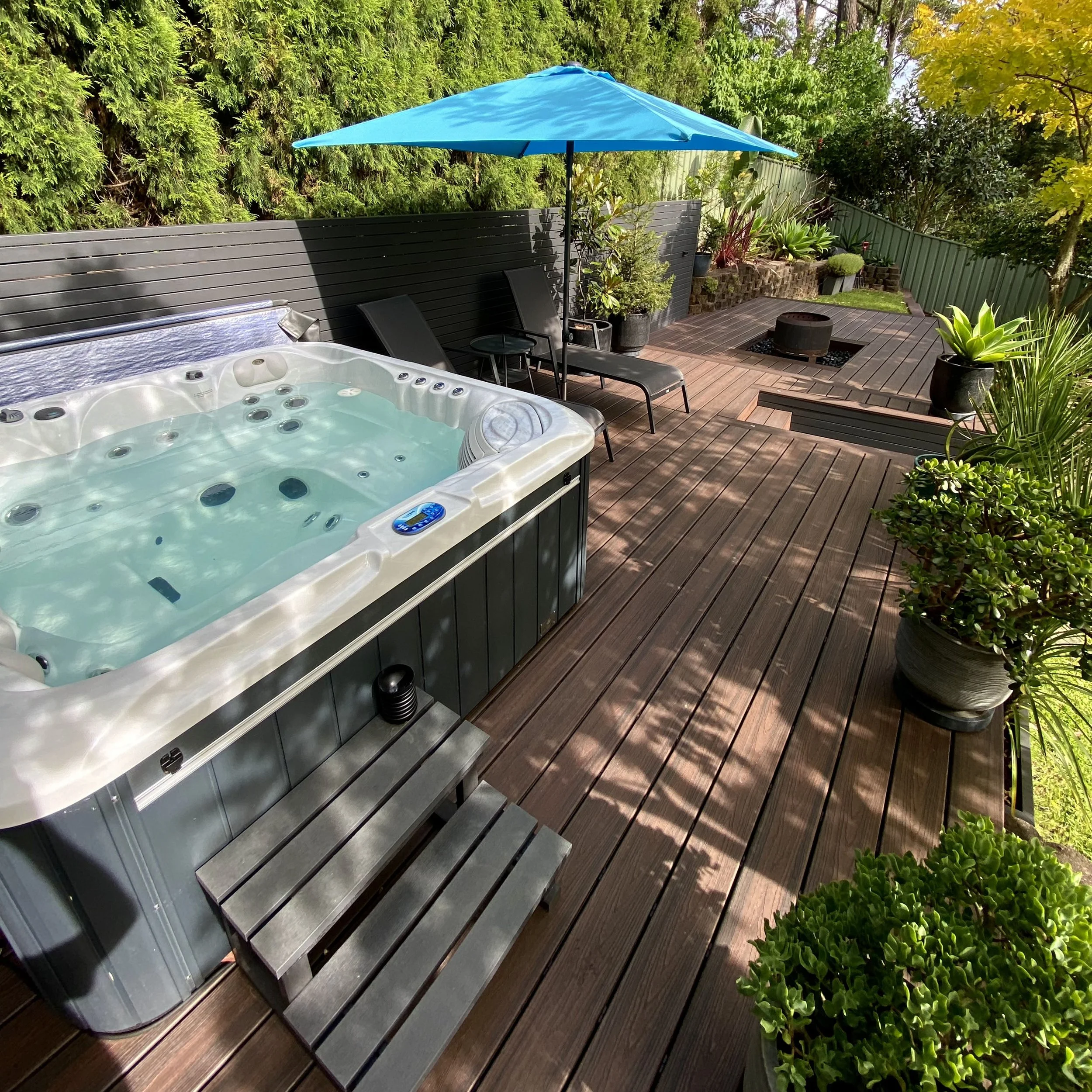 A rich brown composite deck wrapping a cosy outdoor spa surrounded by lush garden touches, crafted by Platinum Decks as part of a warm backyard retreat