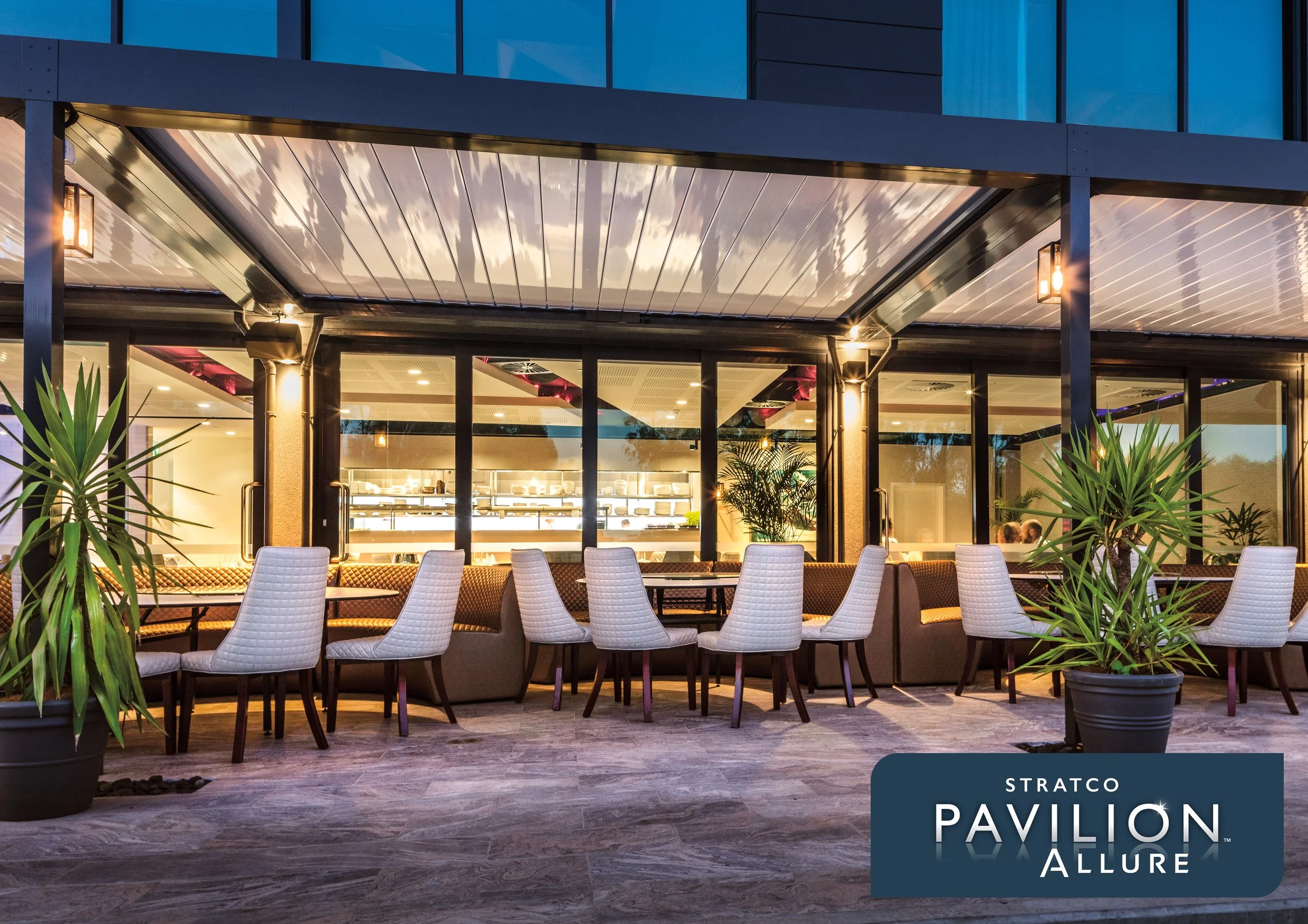 Exterior view of a modern building with large glass windows and a patio and pergola. There are white outdoor chairs arranged around tables and potted plants in the foreground. Platinum Decks authorised installer.