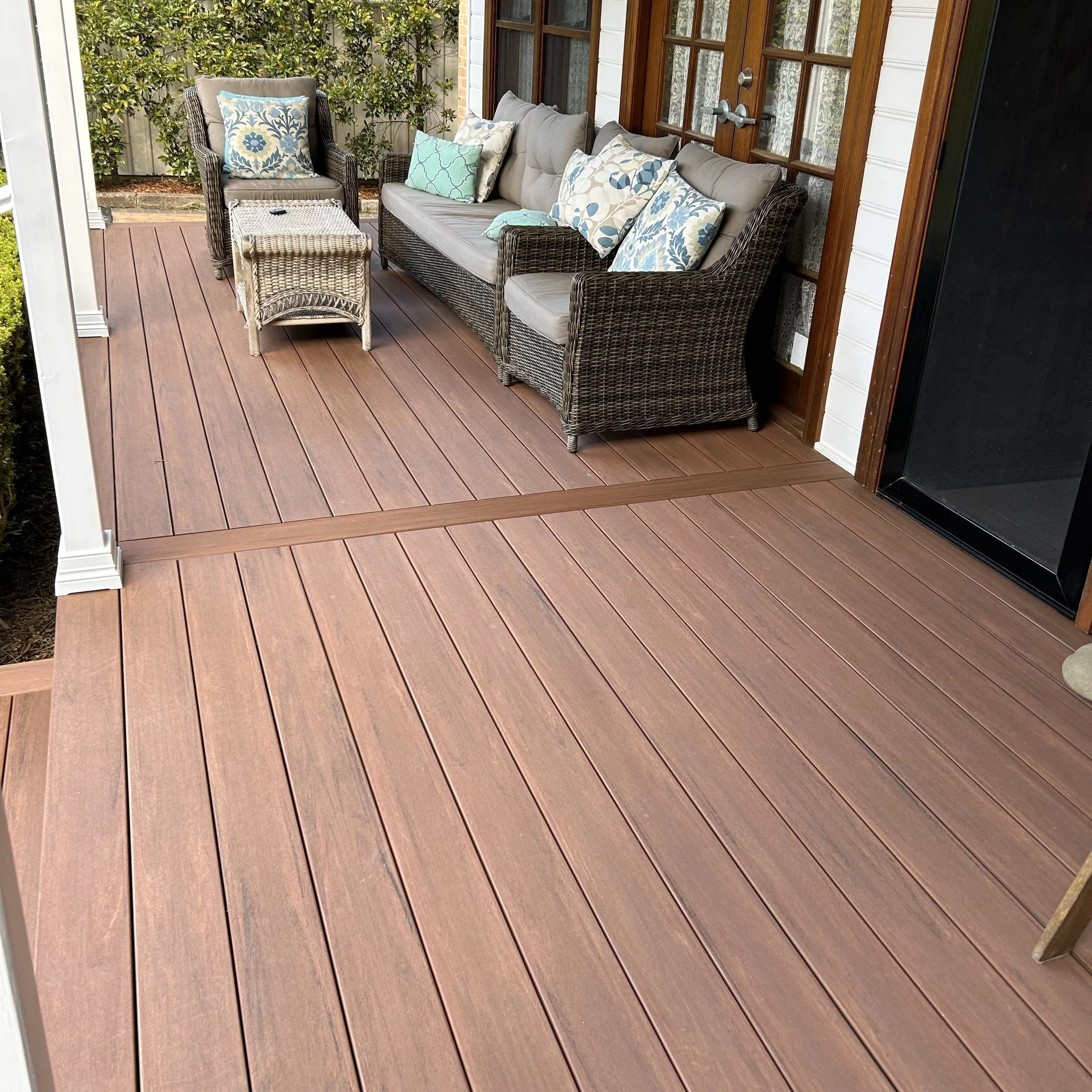 Outdoor new composite deck with wicker furniture and cushions, with a background of bushes and a white house with wooden window frames.