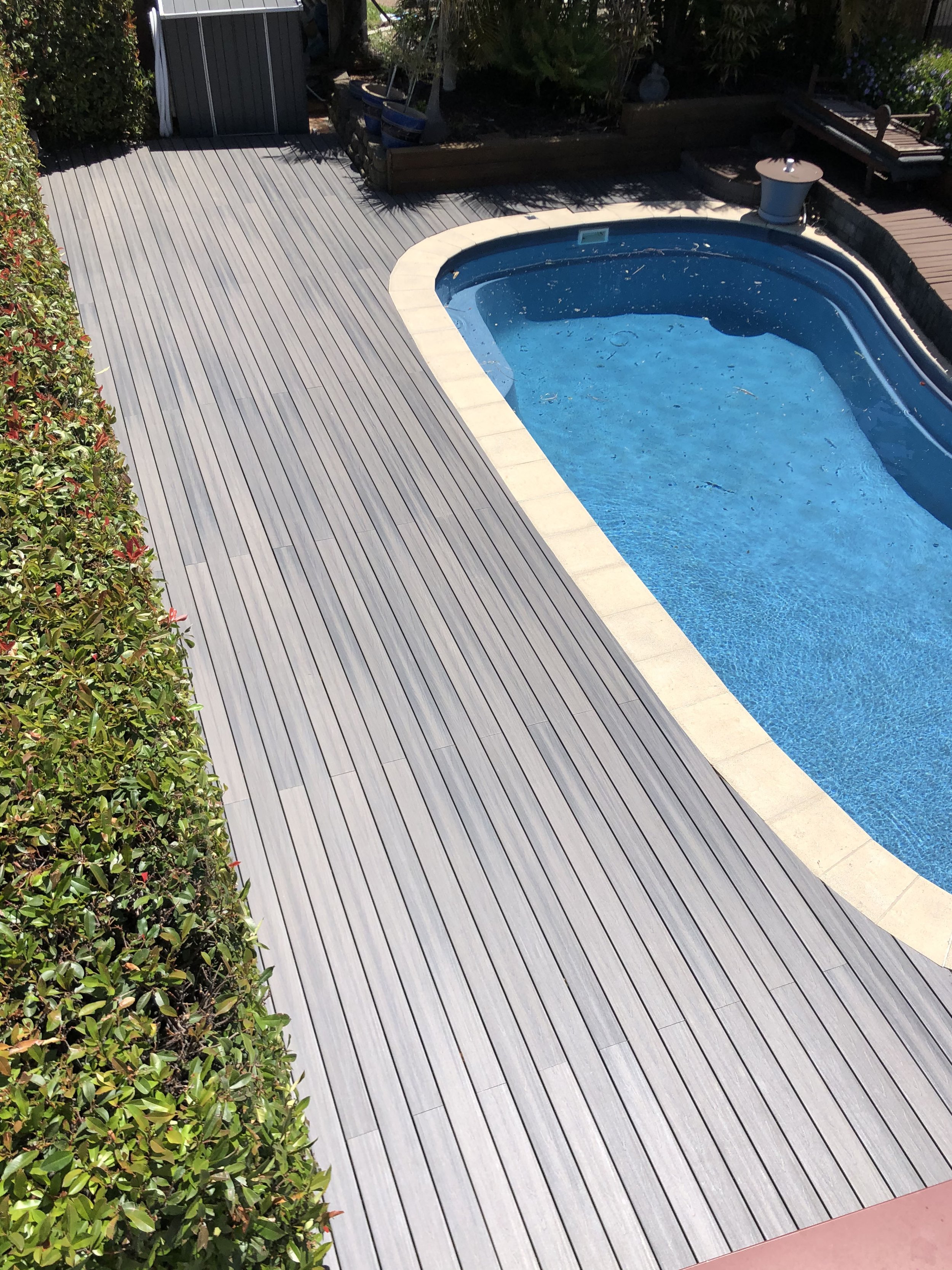 An outdoor scene showcasing a rectangular swimming pool with blue water, surrounded by a light-colored stone border. Adjacent to the pool, there is gray wooden decking extending across the yard. To the left, dense greenery and flowering bushes border