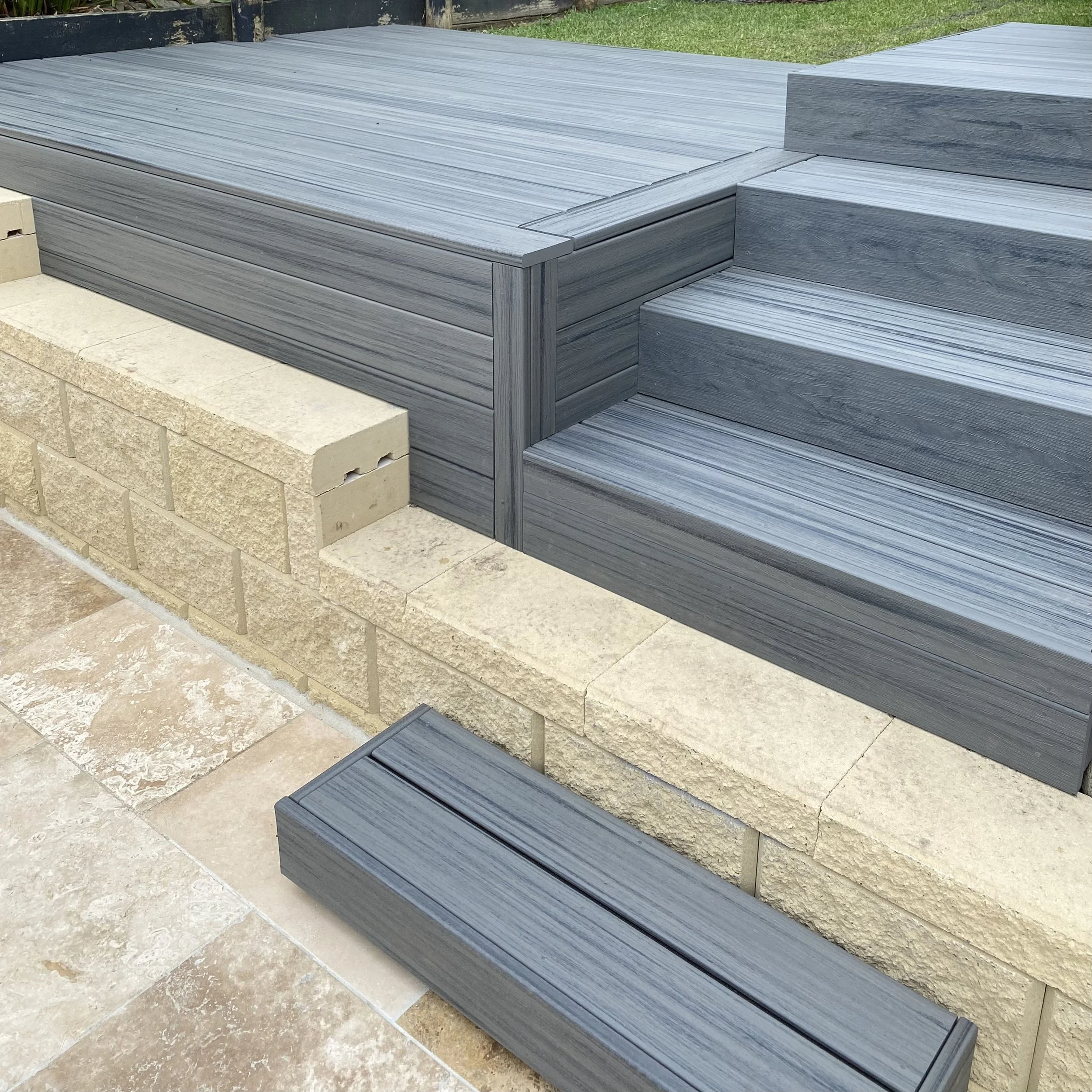 Gray wooden deck with steps, adjacent to a stone patio and a grassy yard.