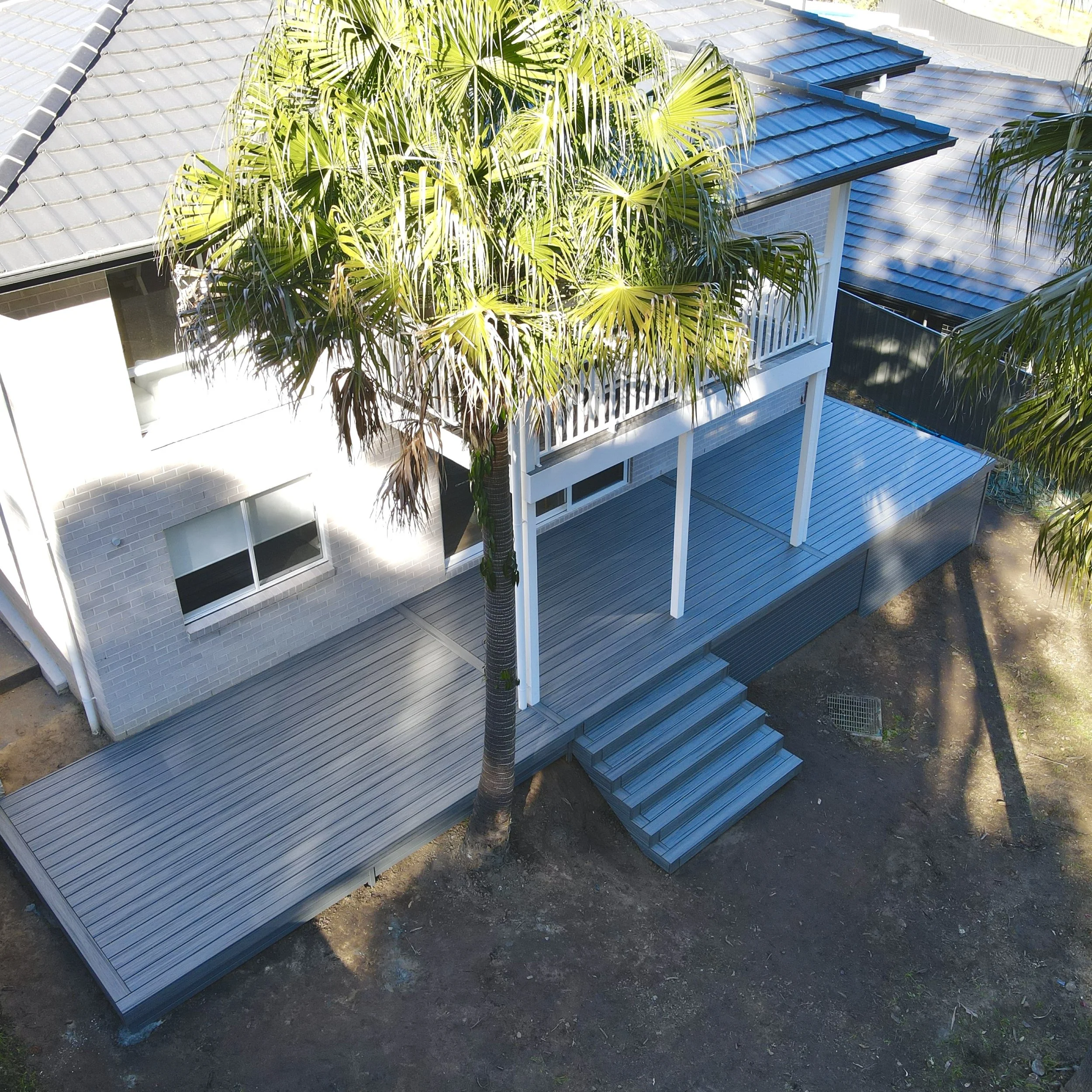 A view of a residential house with a large palm tree in front. The house has a blue metal roof, white balconies, and brick walls. The image is taken from above, showing the house's wooden deck and steps leading to the ground.